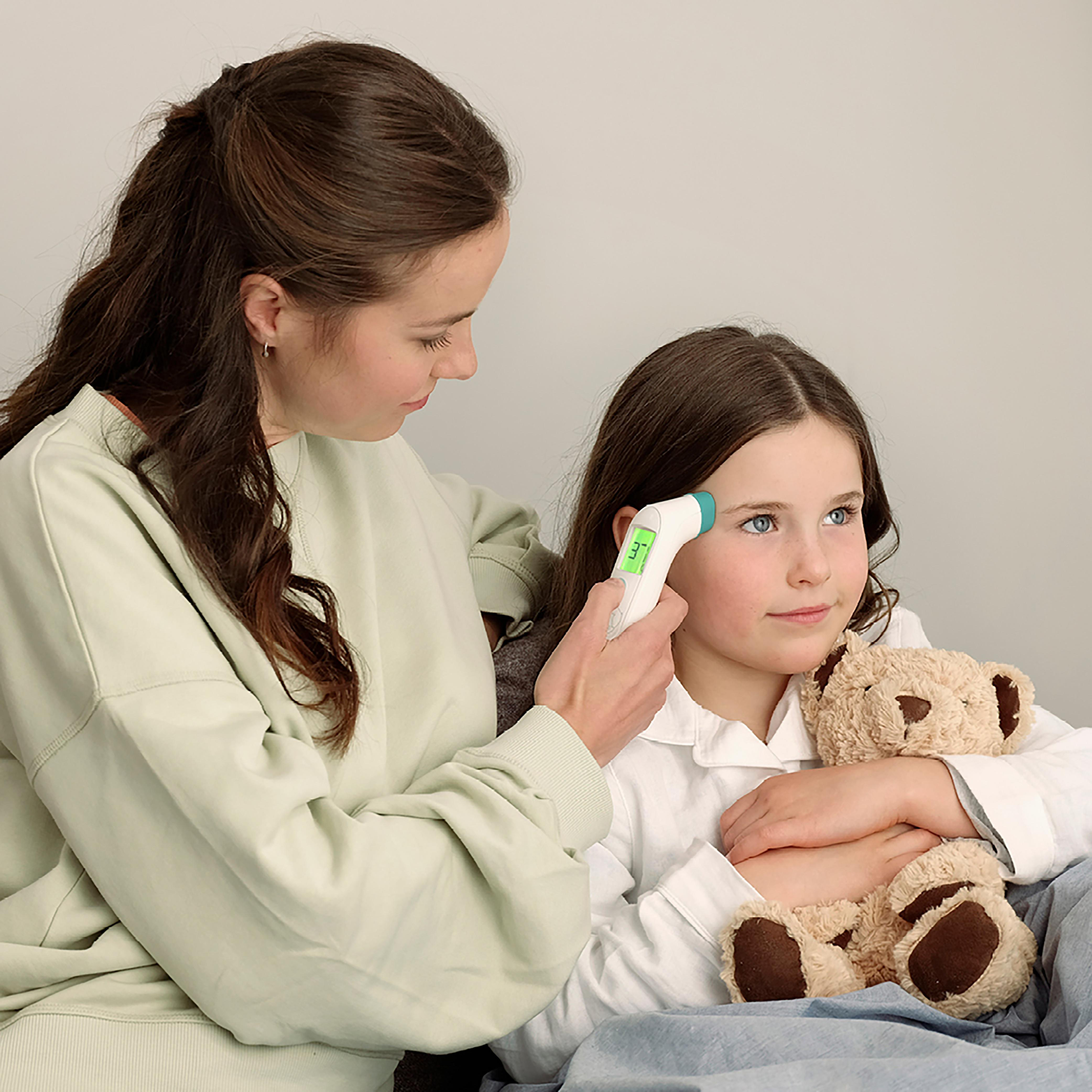 Eine Frau misst die Temperatur eines Kindes mit einem digitalen Thermometer. Ein Teddybär ist in den Armen des Kindes.