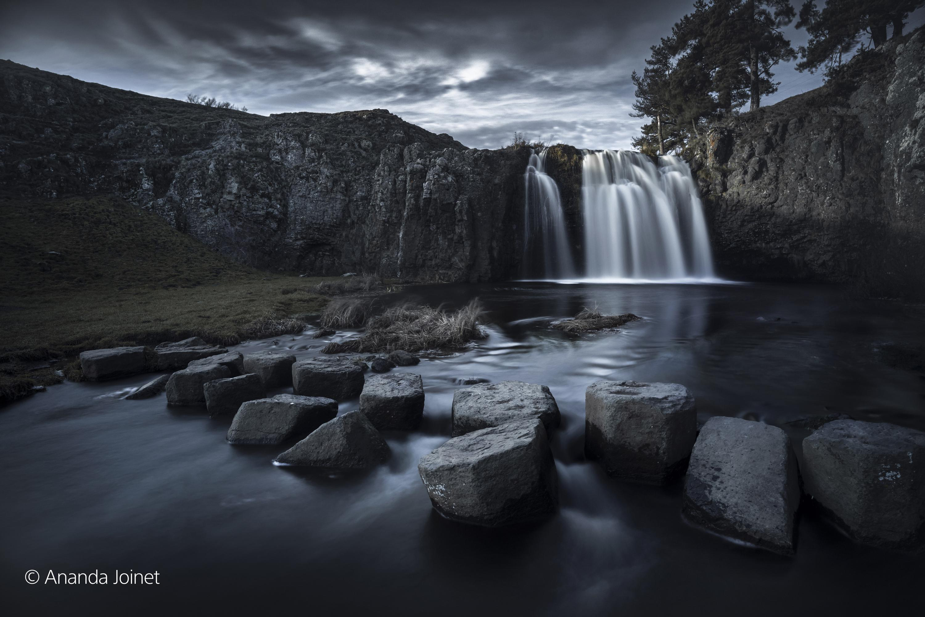 Wasserfall in dunkler, stimmungsvoller Landschaft. Felsen im Vordergrund, wolkiger Himmel. Schwarzweiß.