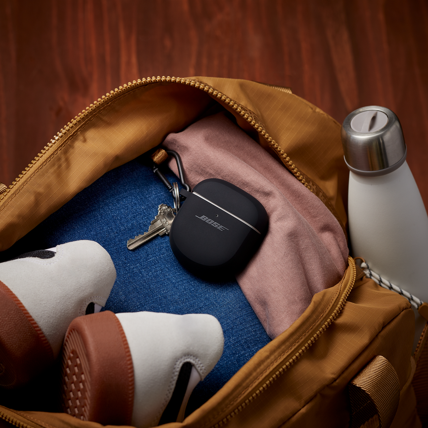 Een zwarte Bose oortelefoon case, sleutels, schoenen en een waterfles in een tas.