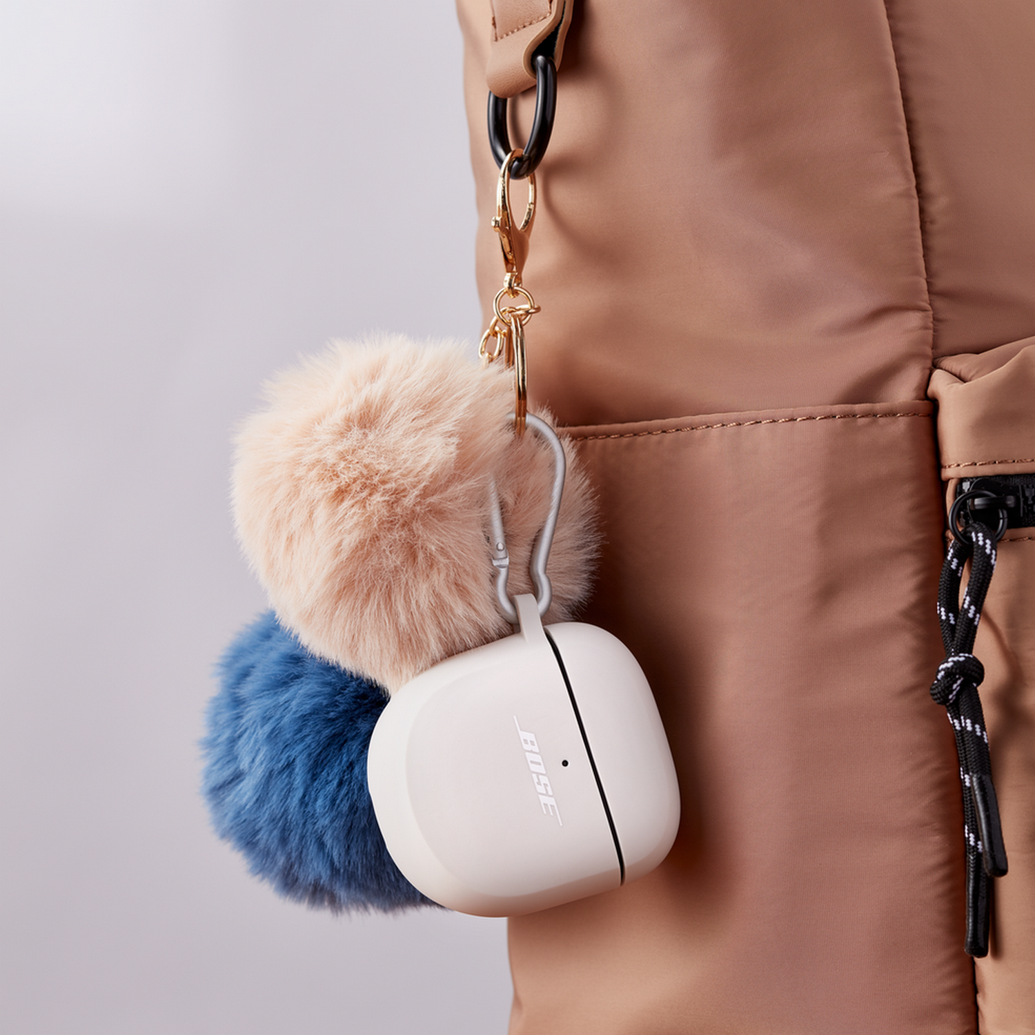Bose oordopjes in een witte case, bevestigd aan een rugzak met een gouden sluiting en fluffy sleutelhangers.