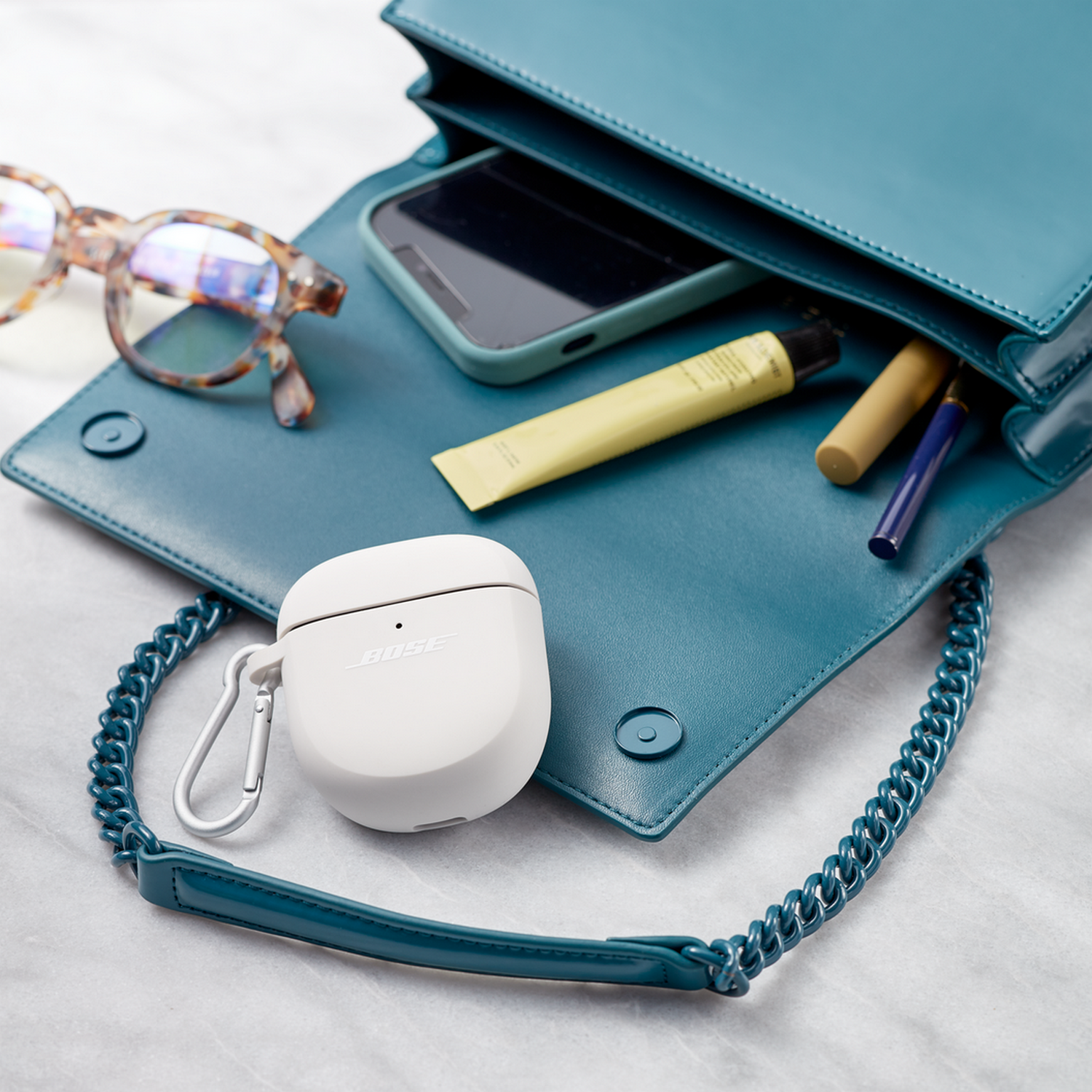 Een turquoise tas met inhoud: bril, telefoon, lippenbalsem en een witte oortelefoonhoes.