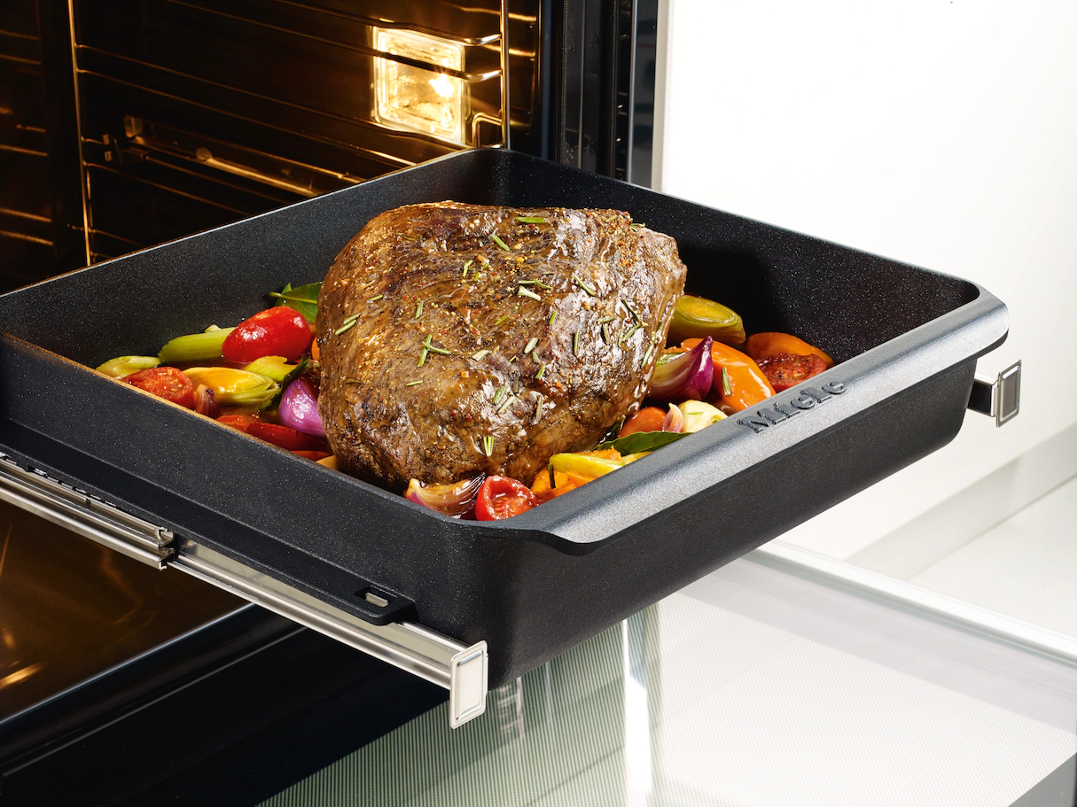 Ein schwarzes Backblech mit gebratenem Fleisch und Gemüse wird aus dem Ofen gezogen.