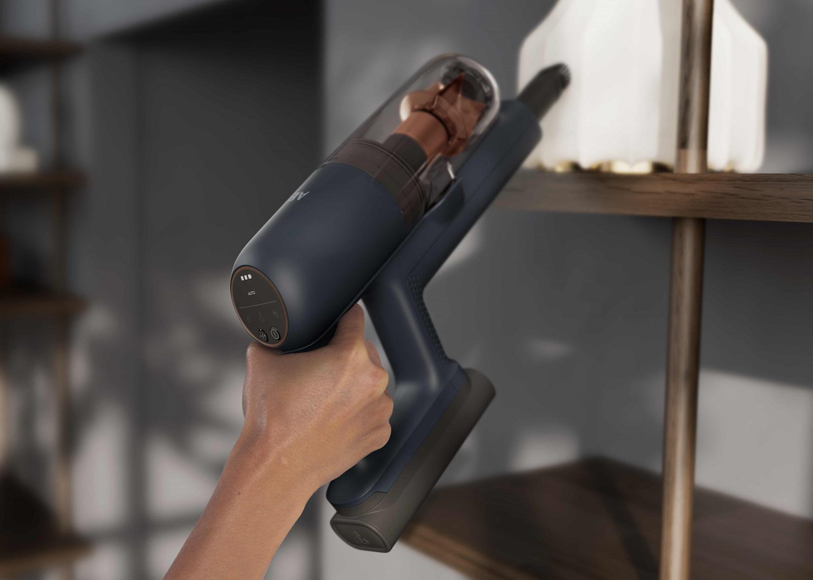Eine Hand hält einen blauen Staubsauger und reinigt ein Regal mit einer weißen Vase. Ein brauner Behälter ist sichtbar.