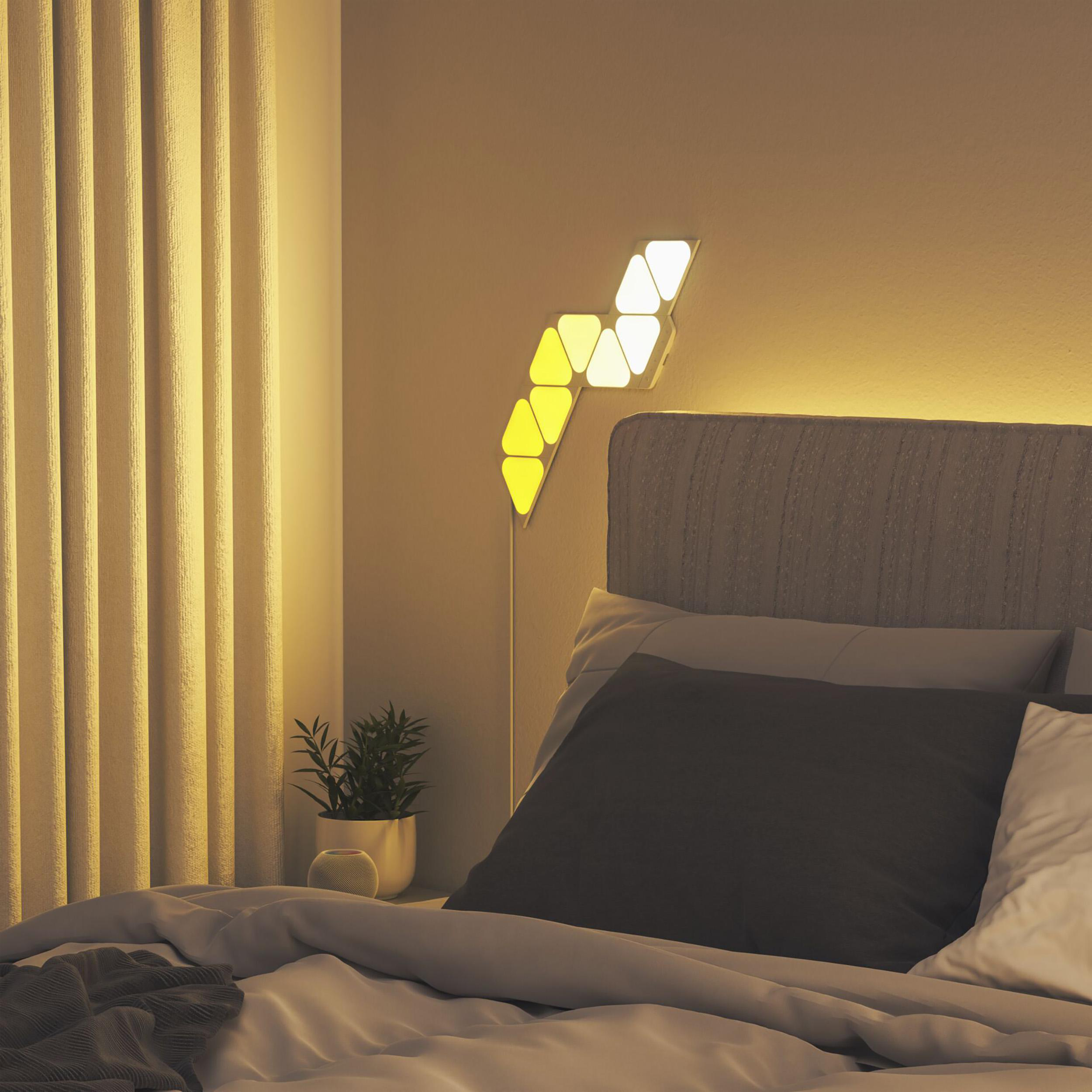 Schlafzimmer mit grauem Bett, moderner Wandleuchte und kleiner Topfpflanze.