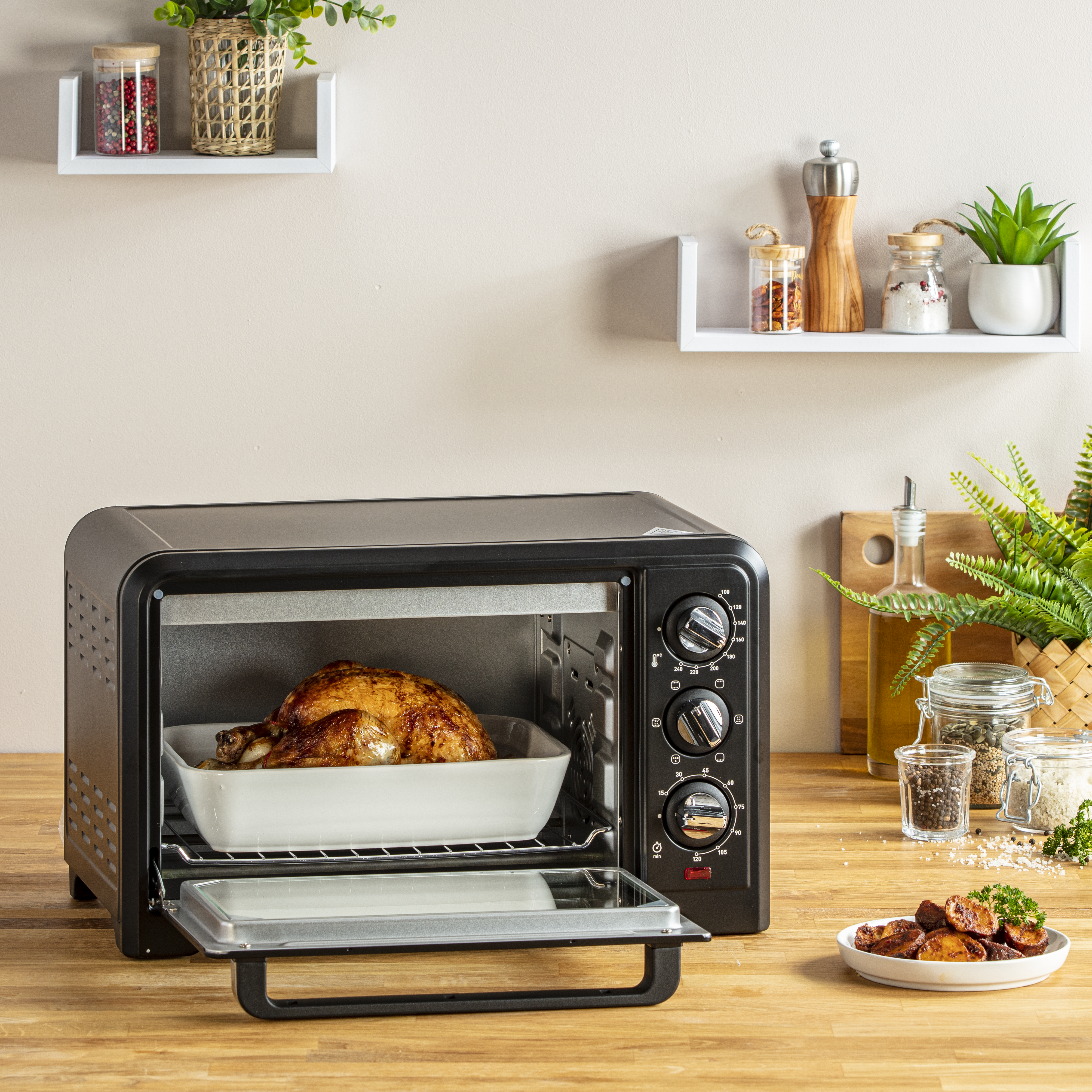 Un horno negro con un pollo asado dentro. Estantes con especias y plantas en el fondo. Plato con comida al frente.