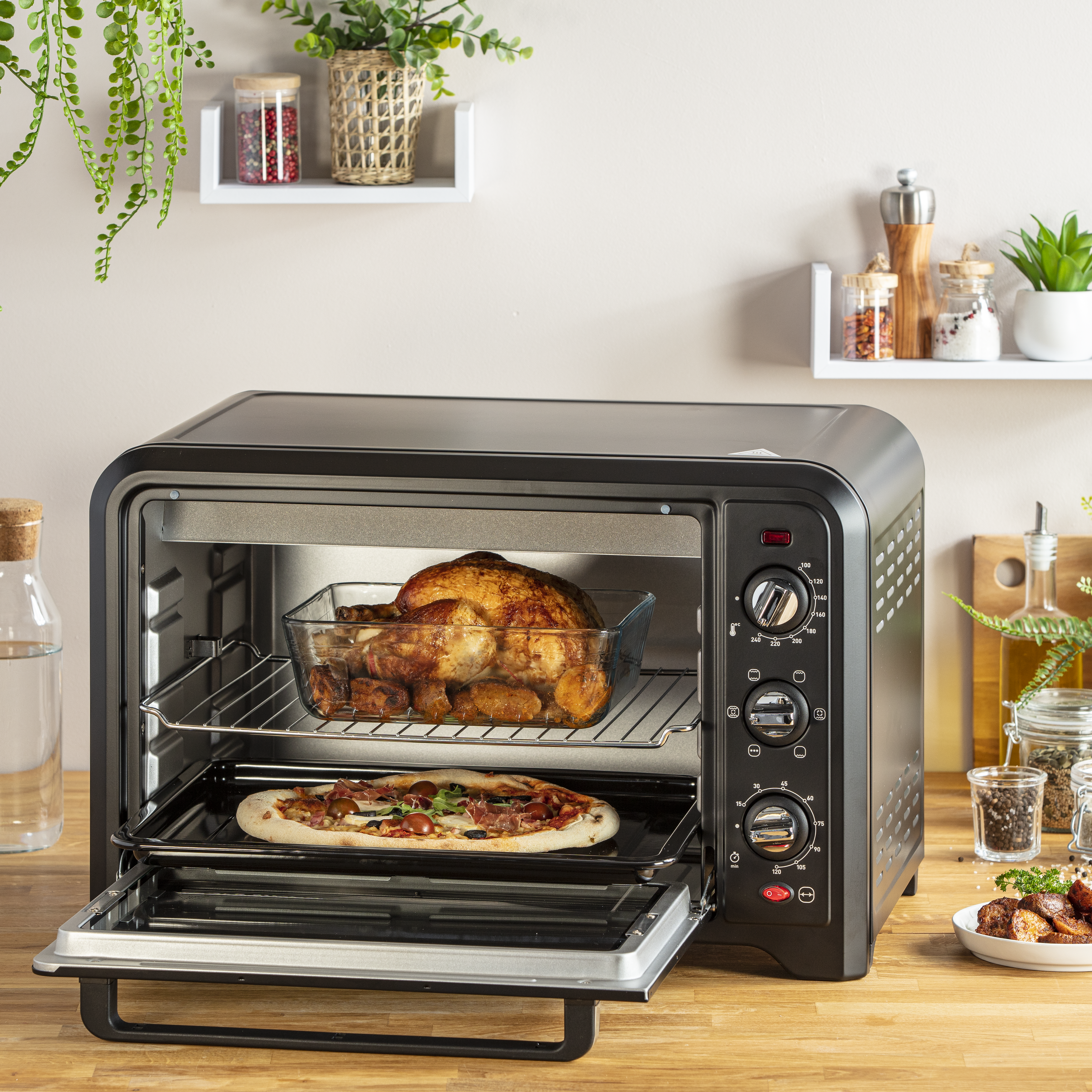 Horno negro con puerta abierta, dentro un pollo asado y una pizza. Encimera de cocina con especias y plantas en el fondo.