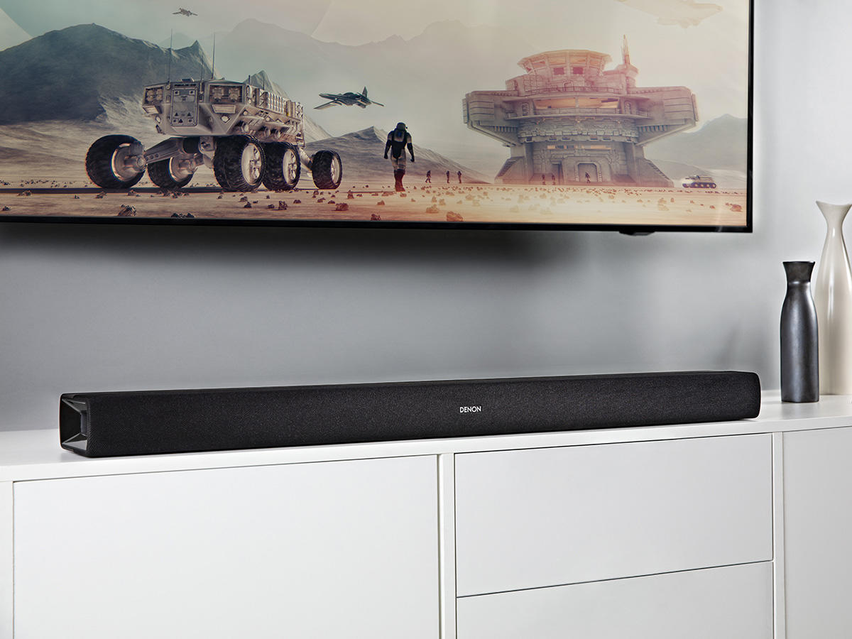 Egy Denon soundbar egy fehér szekrényen egy nagy TV képernyője alatt. A jelenet egy modern nappaliban játszódik.