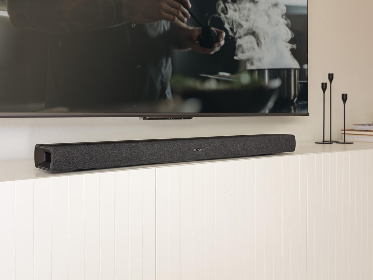 Egy fekete soundbar egy fehér felületen van egy TV alatt.