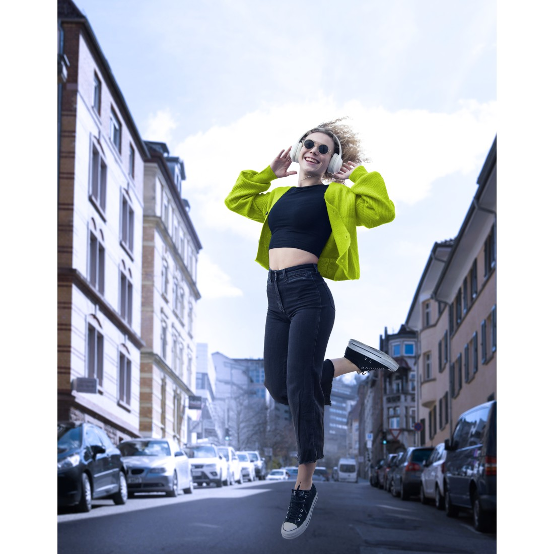 Mujer saltando en la ciudad, con auriculares y gafas. Edificios, cielo soleado y coches al fondo.