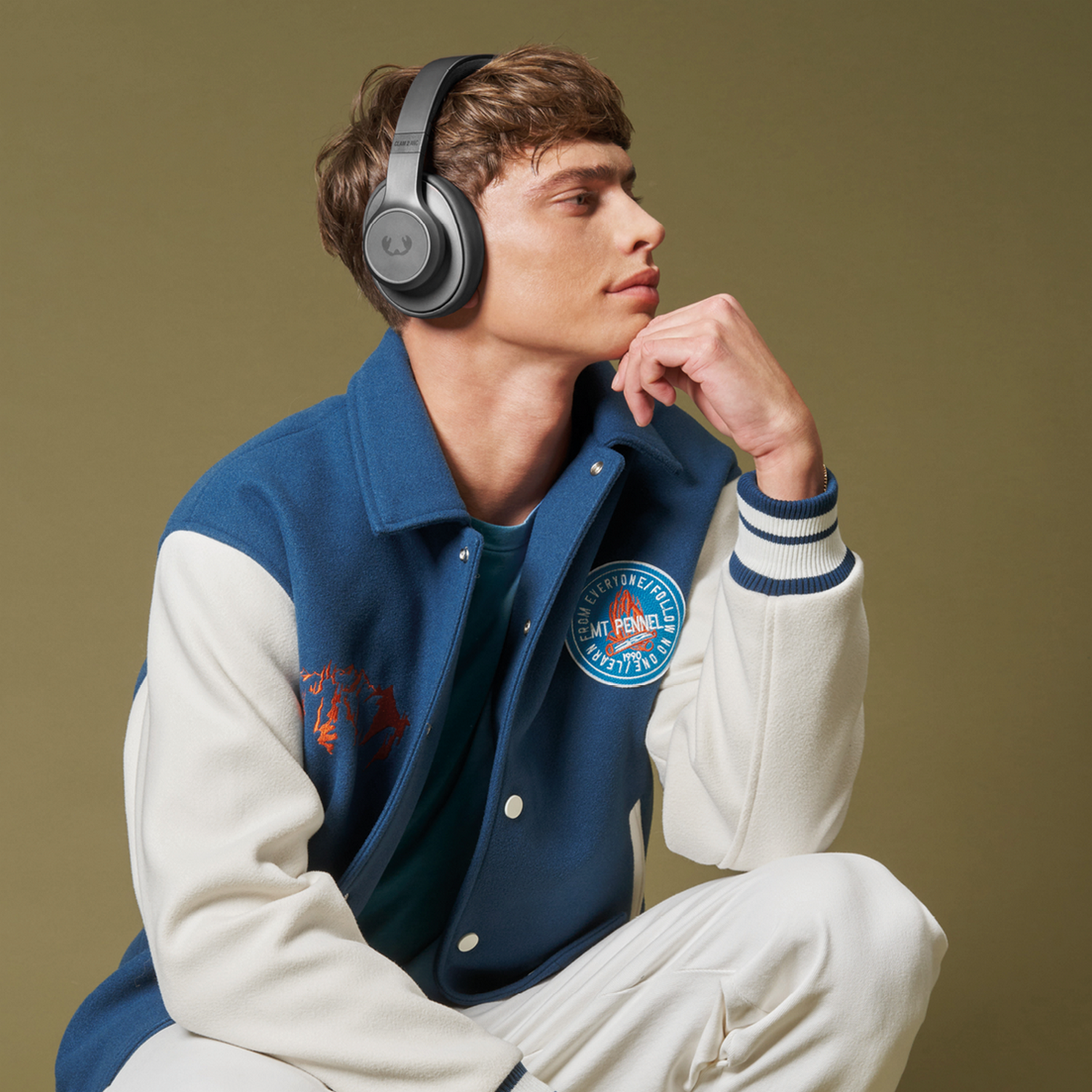 Man met koptelefoon en varsity jacket, kijkt naar rechts. Olijfgroene achtergrond.