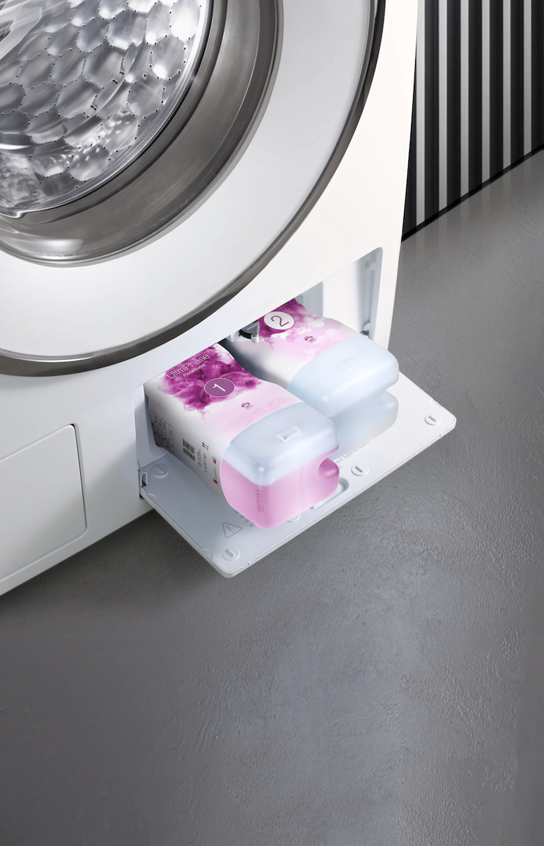 Waschmaschinenschublade offen mit zwei rosa-blauen Waschmittelpackungen.
