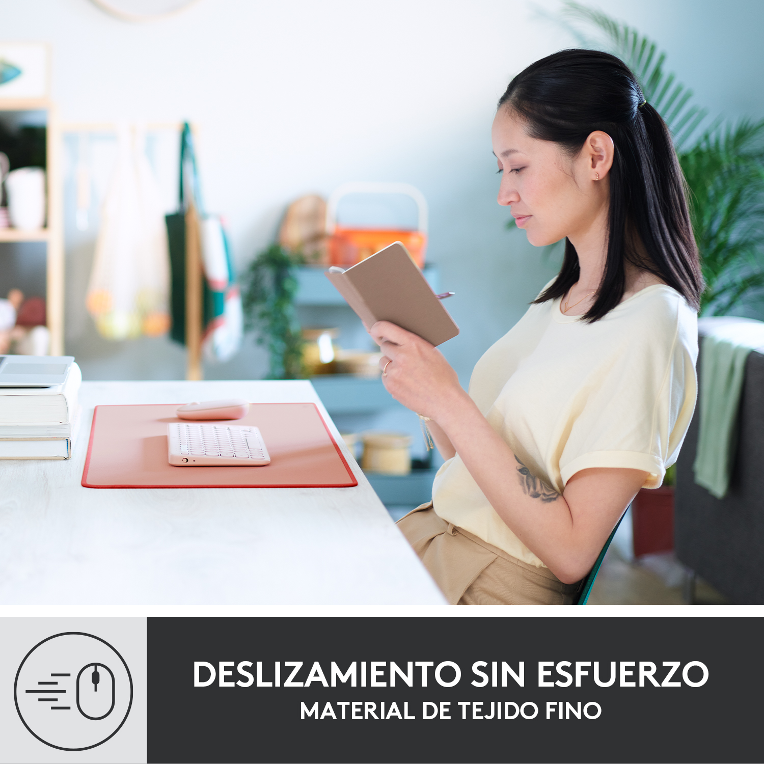 Una mujer escribe en un cuaderno en una mesa con una alfombrilla rosa y un teclado. Una ventana al fondo.