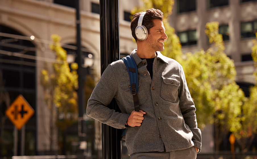 Man with headphones, backpack, walking on a city street. Buildings and trees in the background.