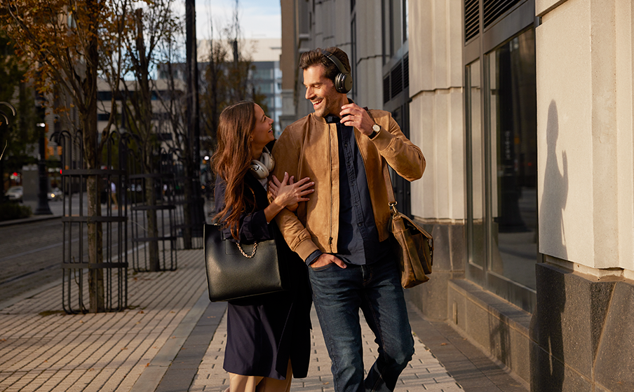 Couple smiling while walking on a sidewalk. Man wears headphones and jacket. Building in the background.