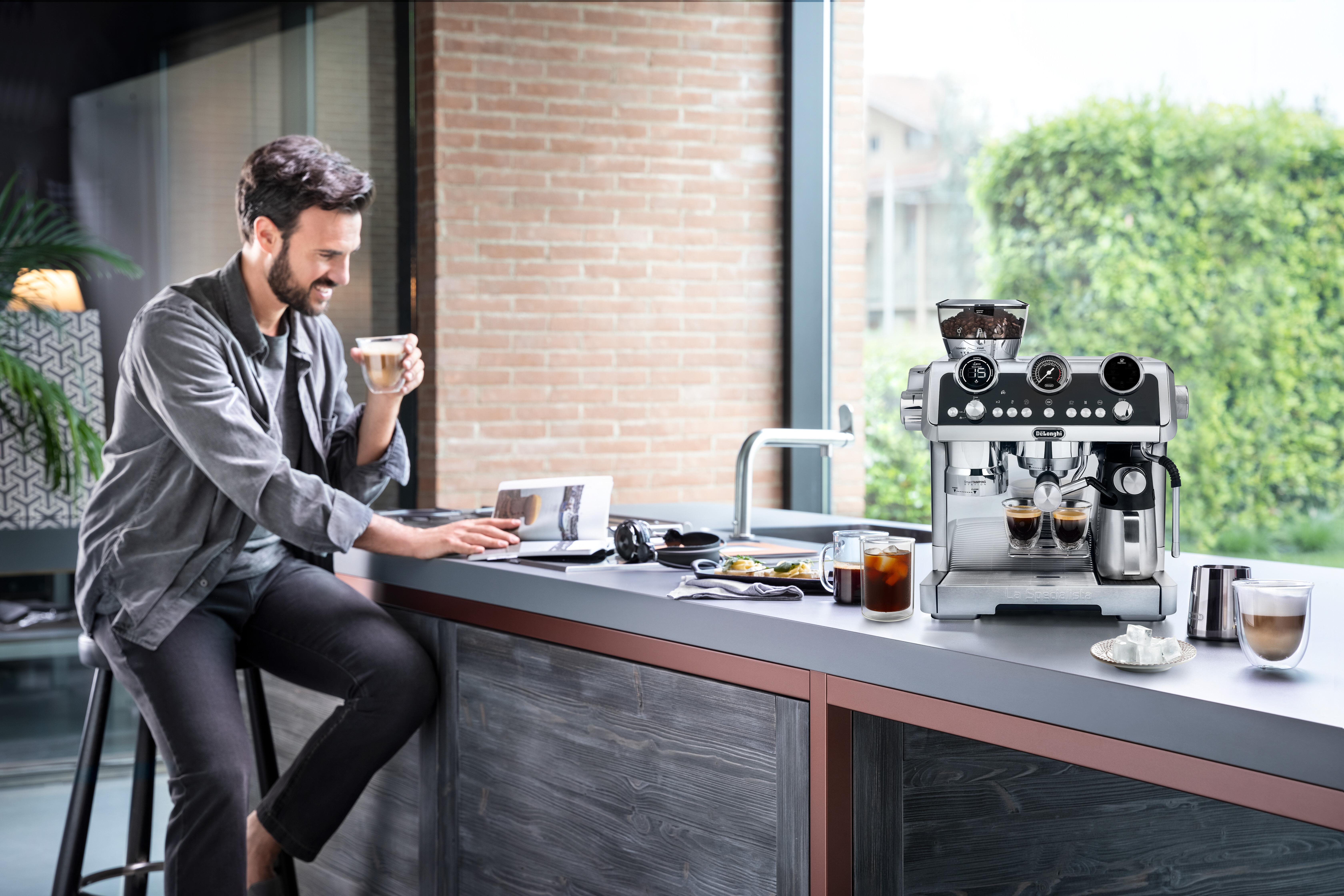 Ein Mann sitzt an einer Theke und benutzt einen Laptop, daneben eine Espressomaschine und Kaffeetassen.