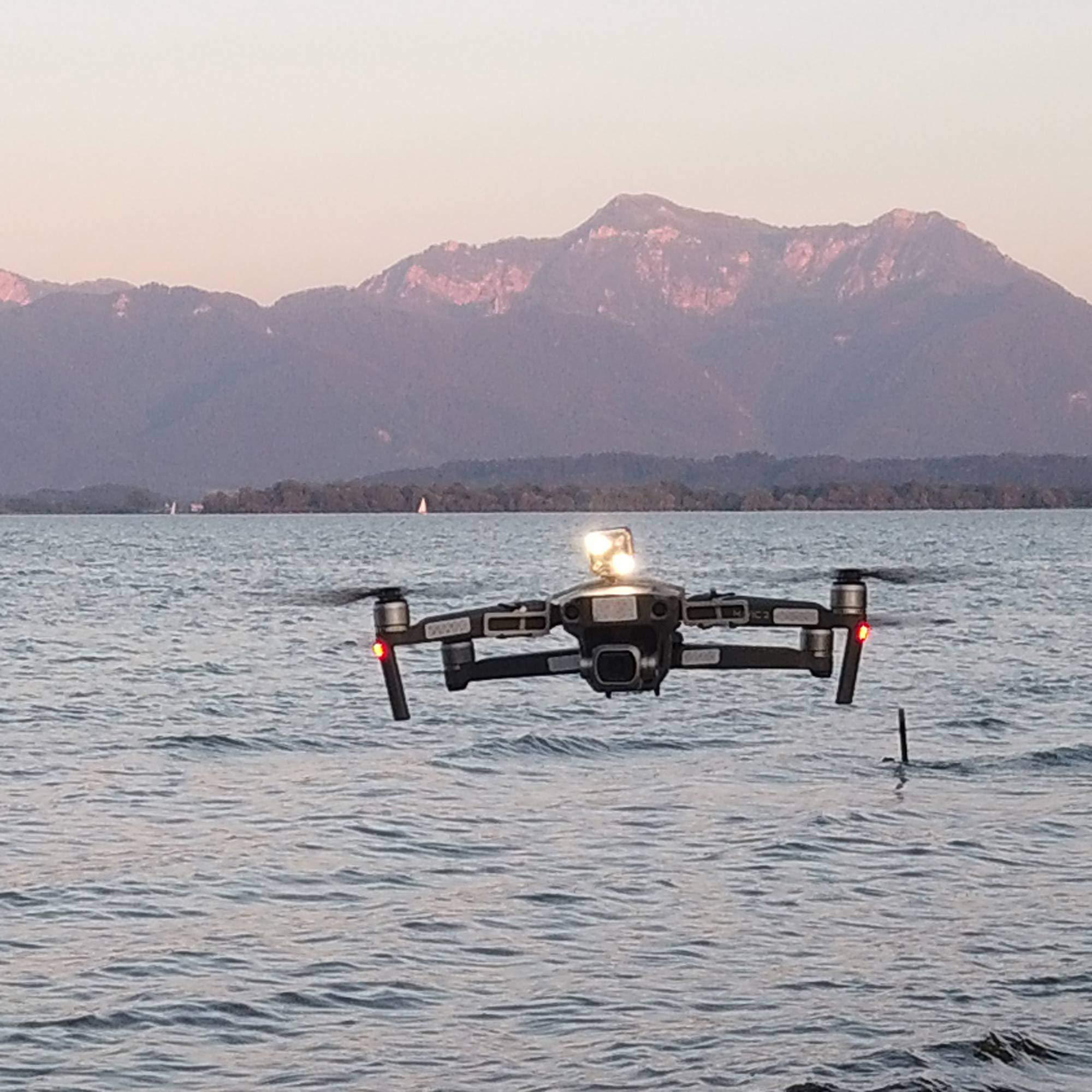 Eine Drohne fliegt über Wasser, Berge im Hintergrund, Sonnenuntergang.