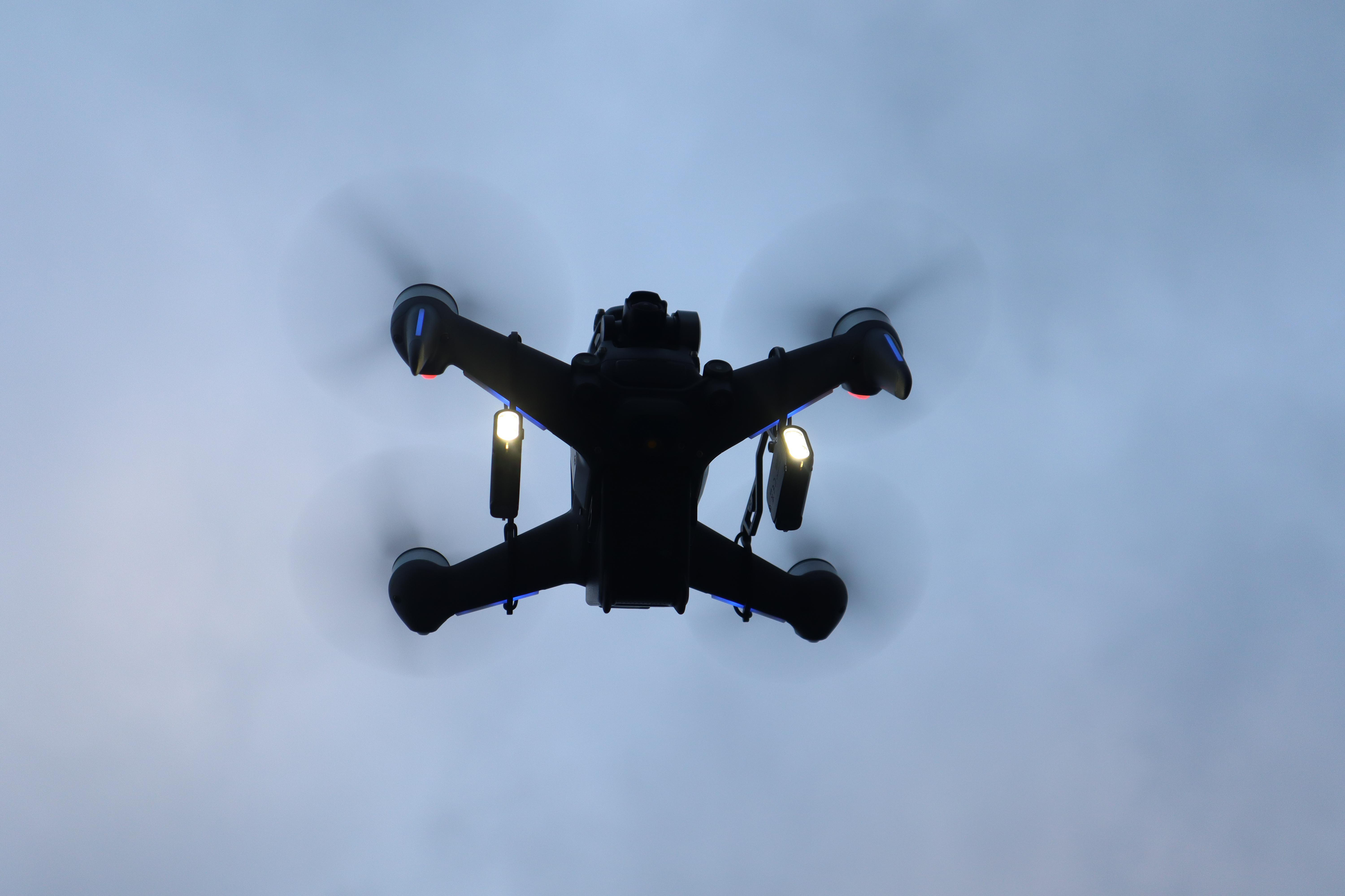Eine schwarze Drohne fliegt am Himmel, ihre Propeller drehen sich, vor grauem Hintergrund.