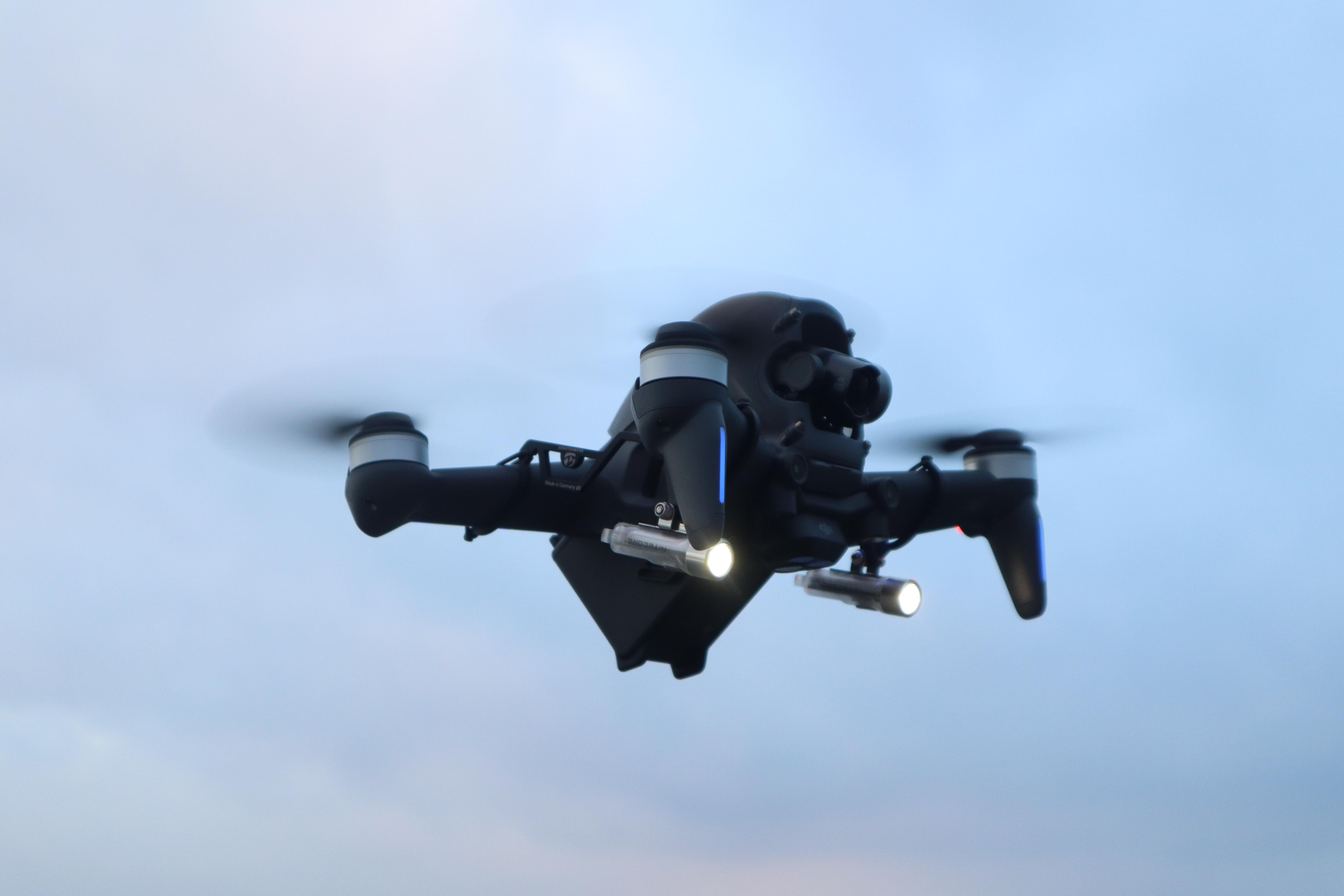 Eine schwarze Drohne mit sich drehenden Propellern fliegt vor einem bewölkten Himmel.