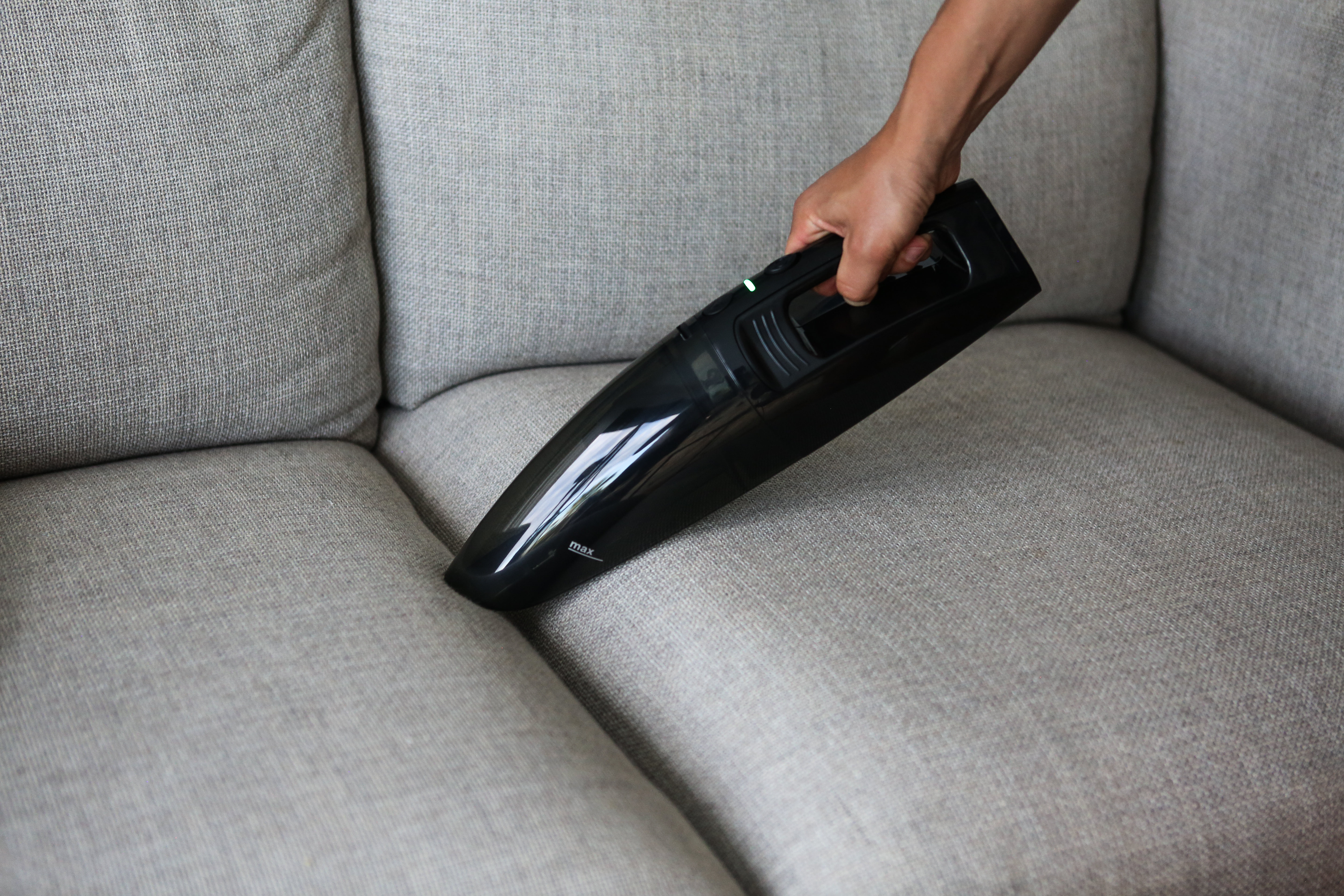 Hand benutzt einen schwarzen Handstaubsauger auf einem grauen Sofa.