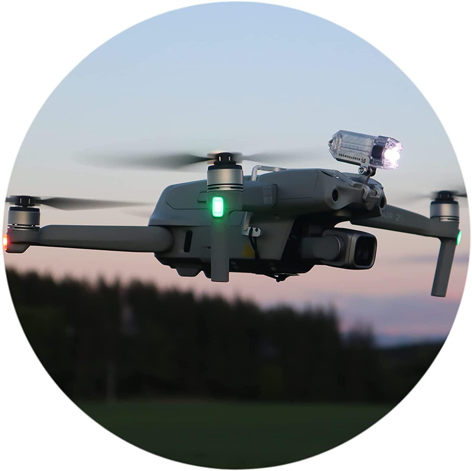 Eine Drohne fliegt in der Luft in der Dämmerung. Die Drohne hat Lichter an. Bäume sind im unscharfen Hintergrund sichtbar.