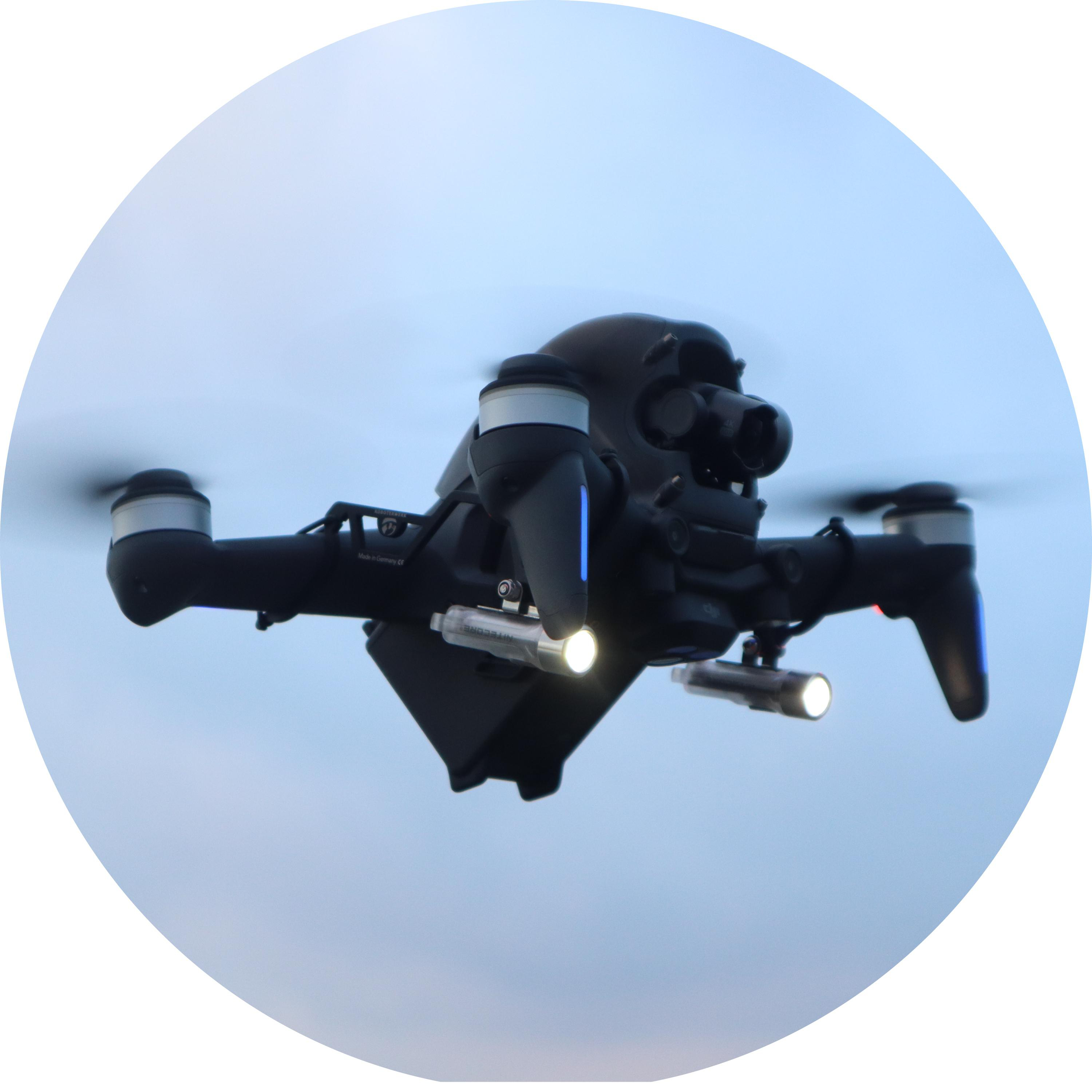 Schwarze Drohne mit Lichtern, die vor einem hellblauen Himmel fliegt.