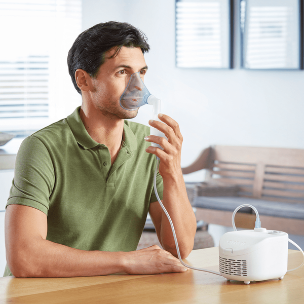 Egy férfi inhalációs maszkot visel. Egy asztalnál ül egy fehér inhalátorral.
