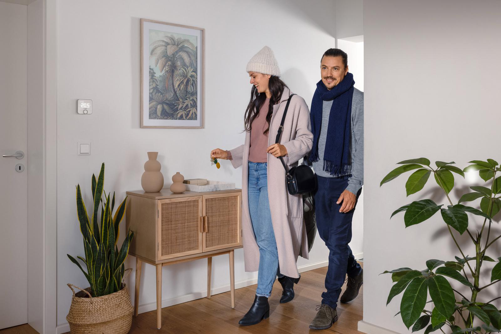 Ein Paar betritt ein Haus. Ein Thermostat, Kunstwerke und Pflanzen sind im Hintergrund sichtbar.