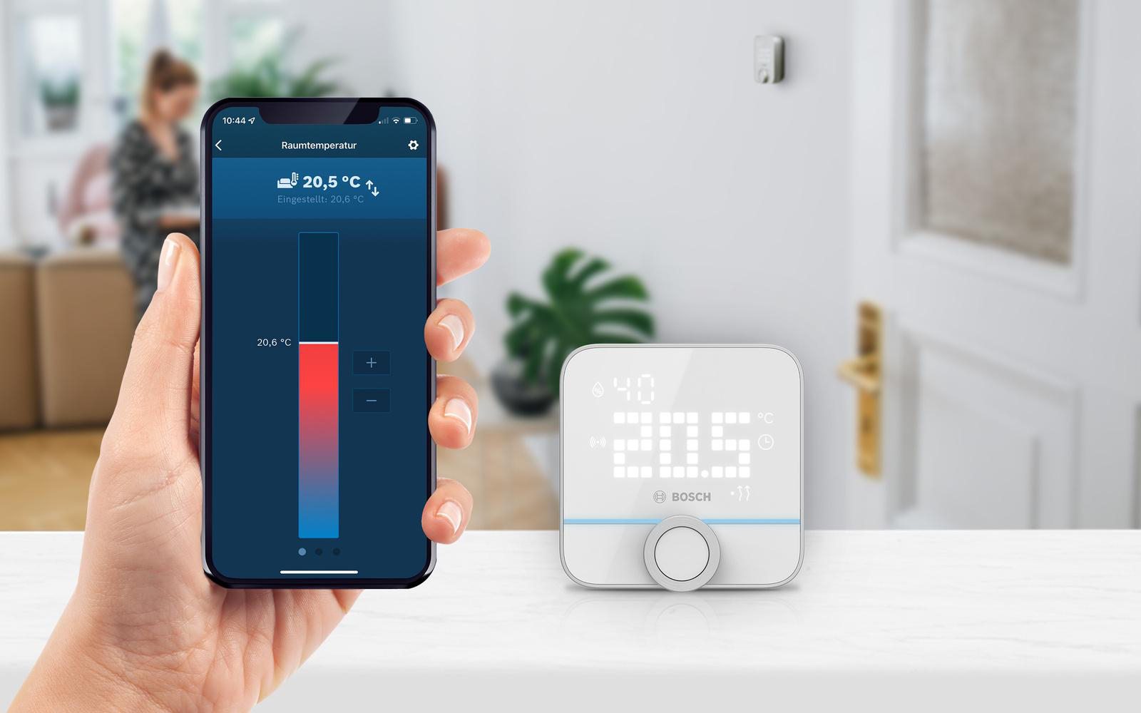 Hand hält Telefon mit Smart-Home-App, neben weißem Thermostat.