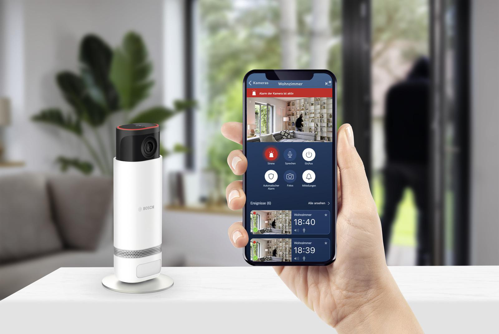 Person hält ein Telefon mit einer Sicherheitskamera-App und eine Kamera auf einem Tisch.