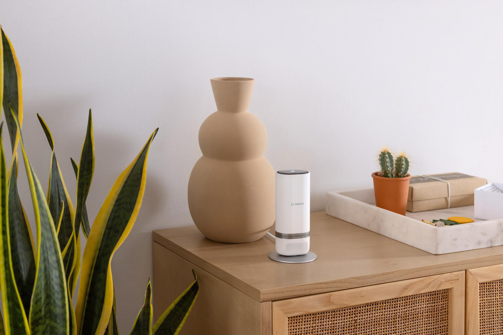 Eine beige Vase und ein weißer Luftreiniger auf einem Holztisch. Eine Pflanze und ein kleiner Kaktus sind auch vorhanden.