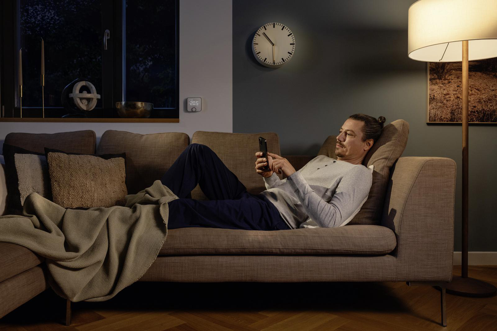 Mann auf Sofa schaut auf Telefon, Uhr und Lampe sichtbar.