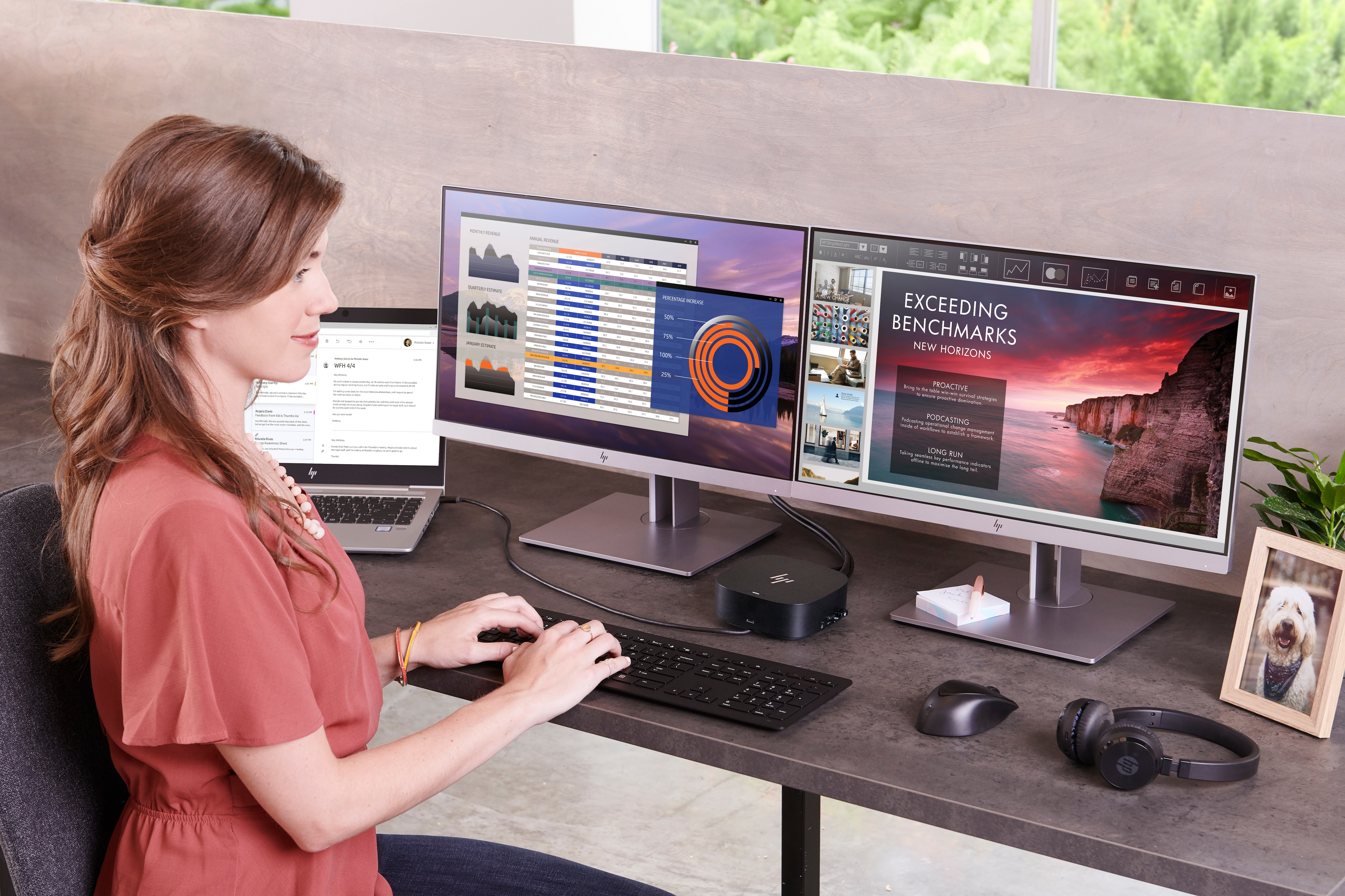 Frau am Schreibtisch mit Laptop und Tastatur, mit Dual-Monitoren, die Daten anzeigen. Ein Hundefoto und Kopfhörer sind vorhanden.