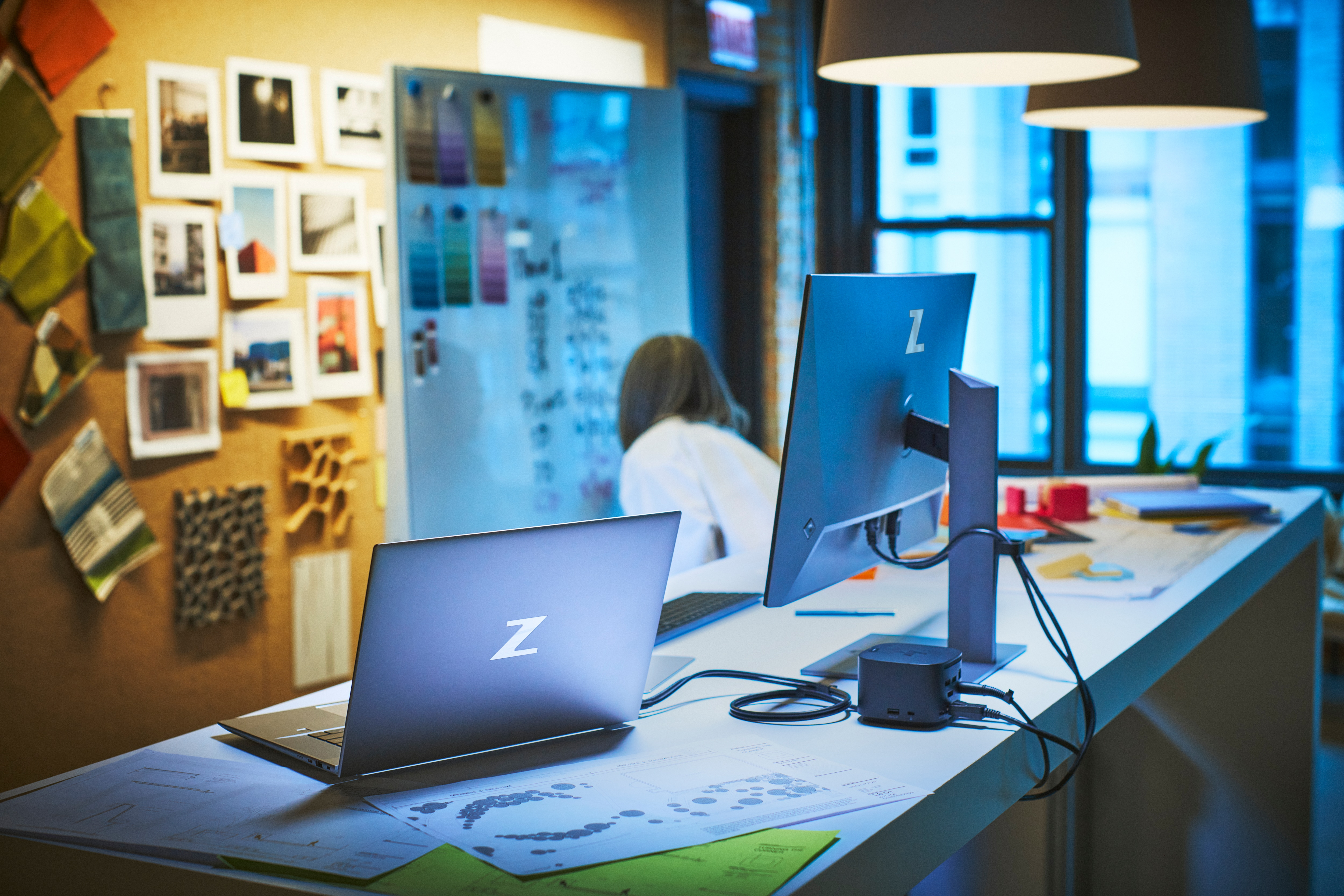 Office with laptop, monitor, and a person working in the background, with a whiteboard.