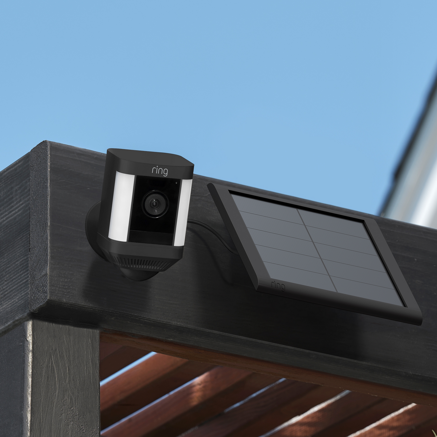 Black security camera with solar panel mounted on a dark wooden structure. Blue sky background.