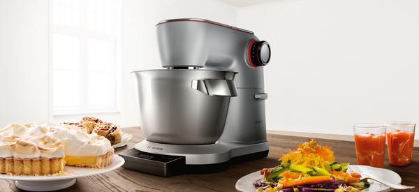 Ein silberner Küchenmixer auf einem Tisch mit Speisen und Getränken an den Seiten.