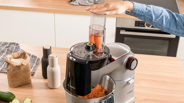 Eine Hand einer Frau gießt Flüssigkeit in eine Küchenmaschine mit geriebenen Karotten.