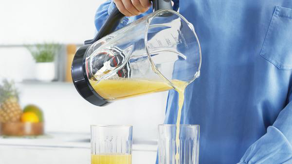 Person gießt gelbe Flüssigkeit aus einem durchsichtigen Krug in Gläser. Heller Küchenhintergrund.