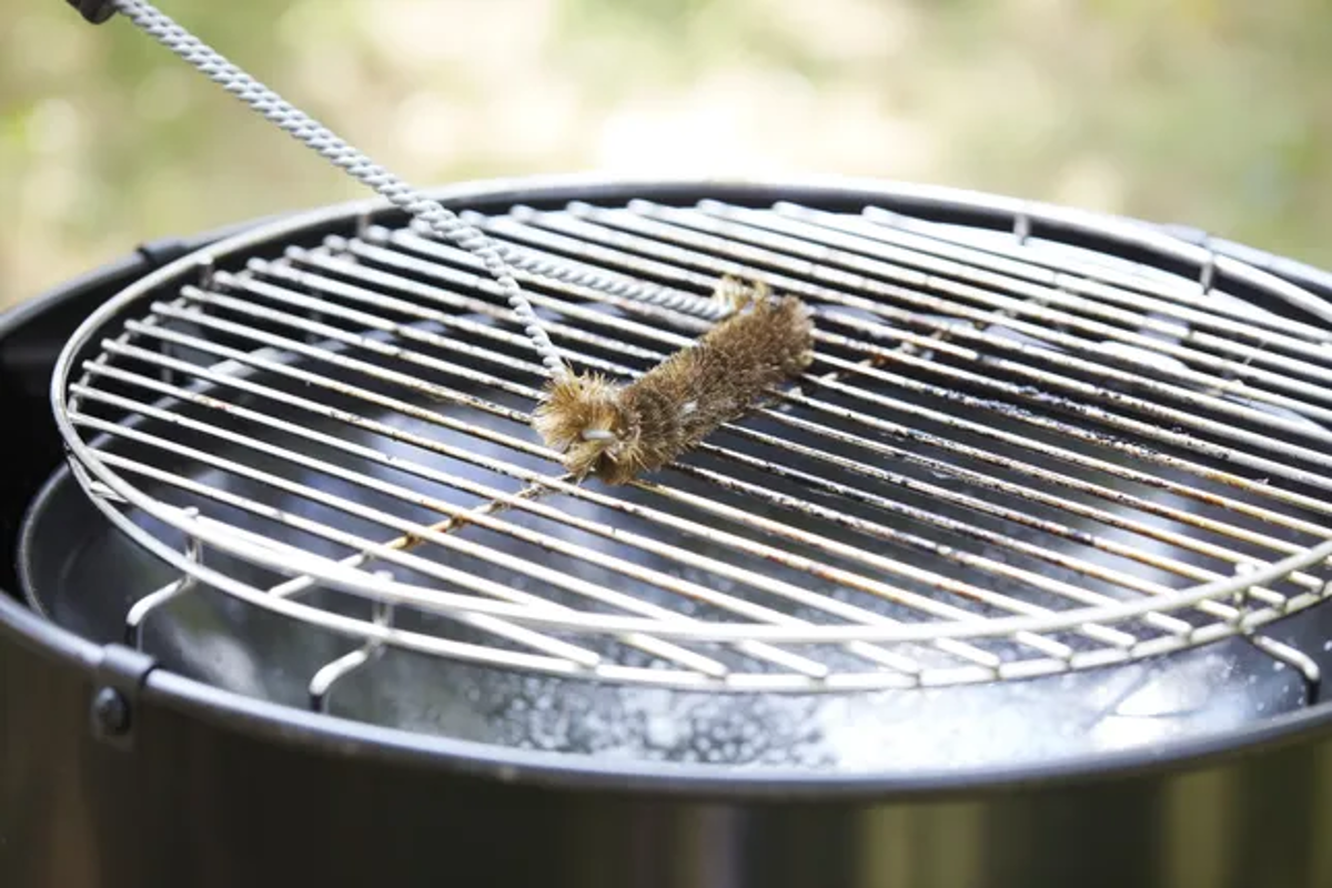 Ein Grillrost wird mit einer Bürste gereinigt. Der Rost befindet sich über einem schwarzen Grill.