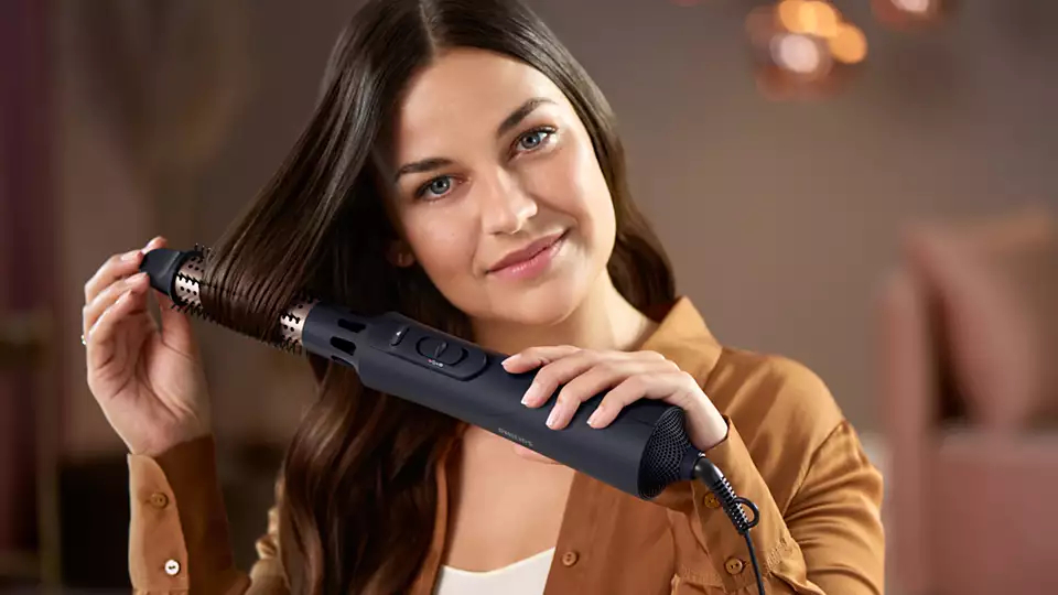 Frau, die ihre Haare mit einem Haartrockner stylt. Sie lächelt und hält den Trockner.