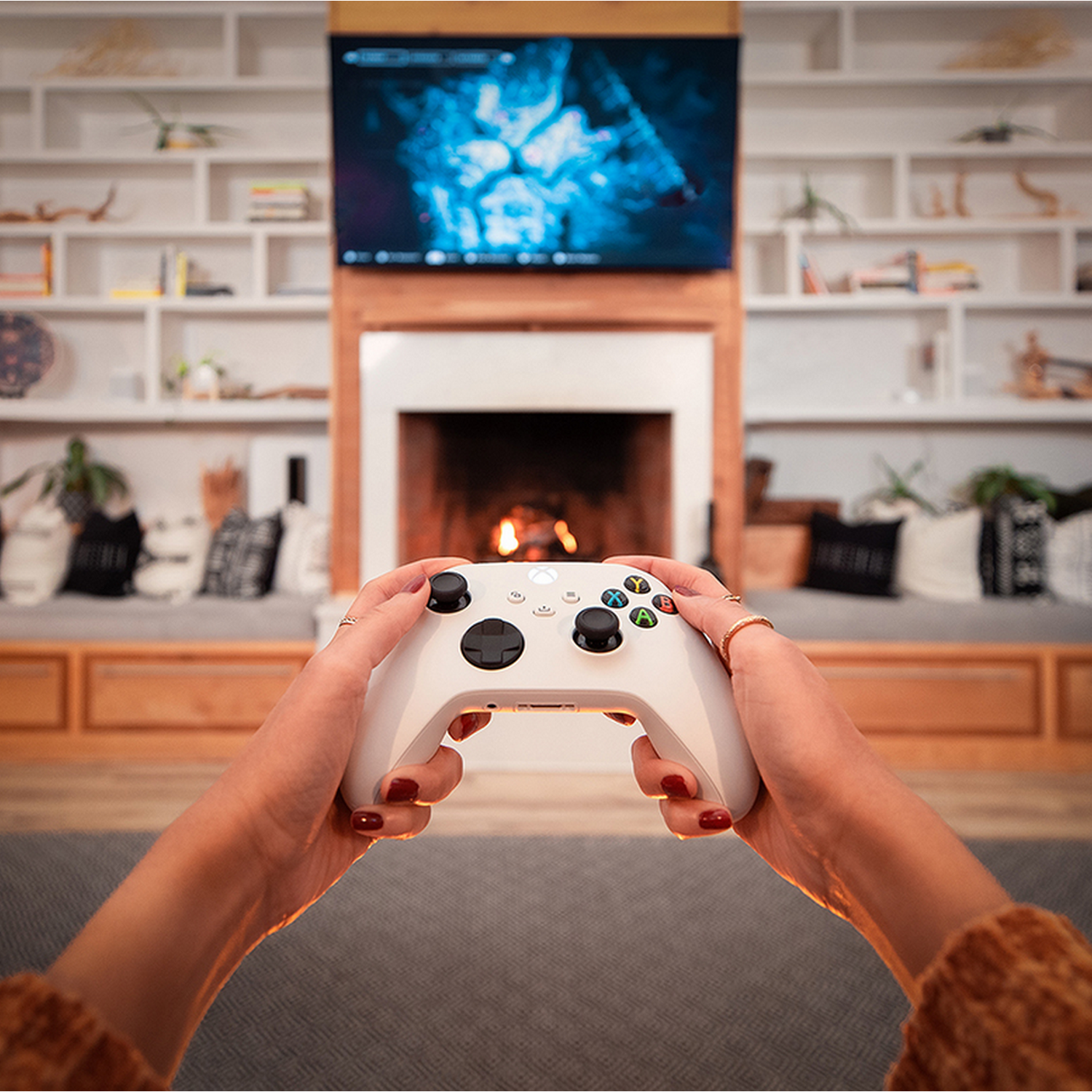 Handen houden witte gamecontroller vast. Tv-scherm toont game. Open haard op achtergrond met planken en kussens.