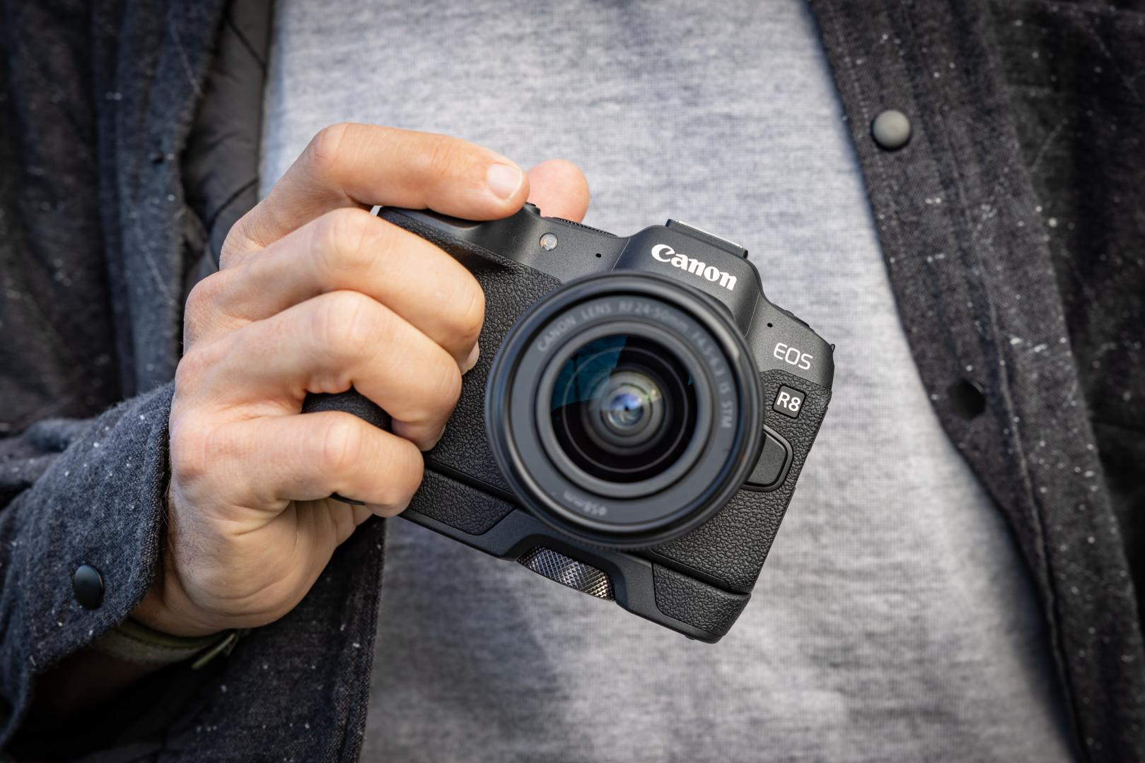 Eine Person hält eine schwarze Canon-Kamera mit einem Objektiv. Graue Jacke im Hintergrund.