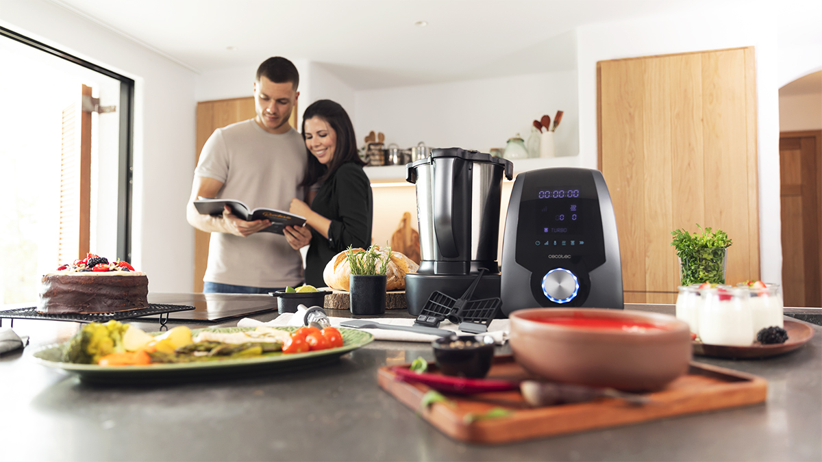 Una pareja en la cocina mirando un libro de cocina. Un procesador de alimentos y alimentos en la encimera.