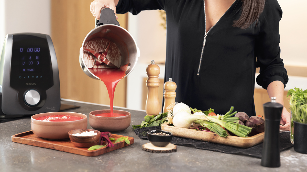 Mujer vertiendo sopa roja de una licuadora en cuencos con ingredientes en un mostrador.