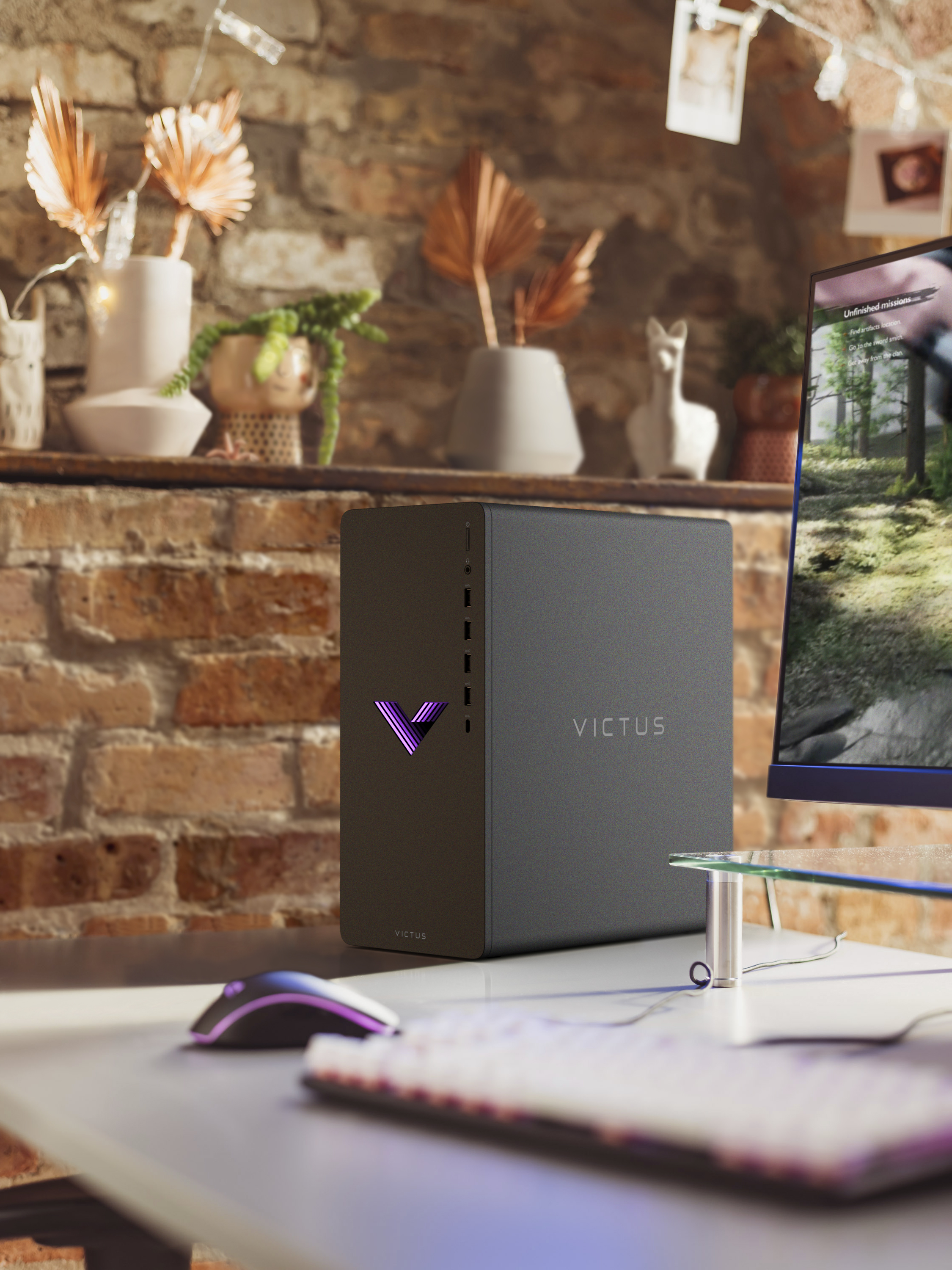 Ein schwarzer Victus Gaming-PC auf einem Schreibtisch, mit Maus und Tastatur. Backsteinmauer im Hintergrund.