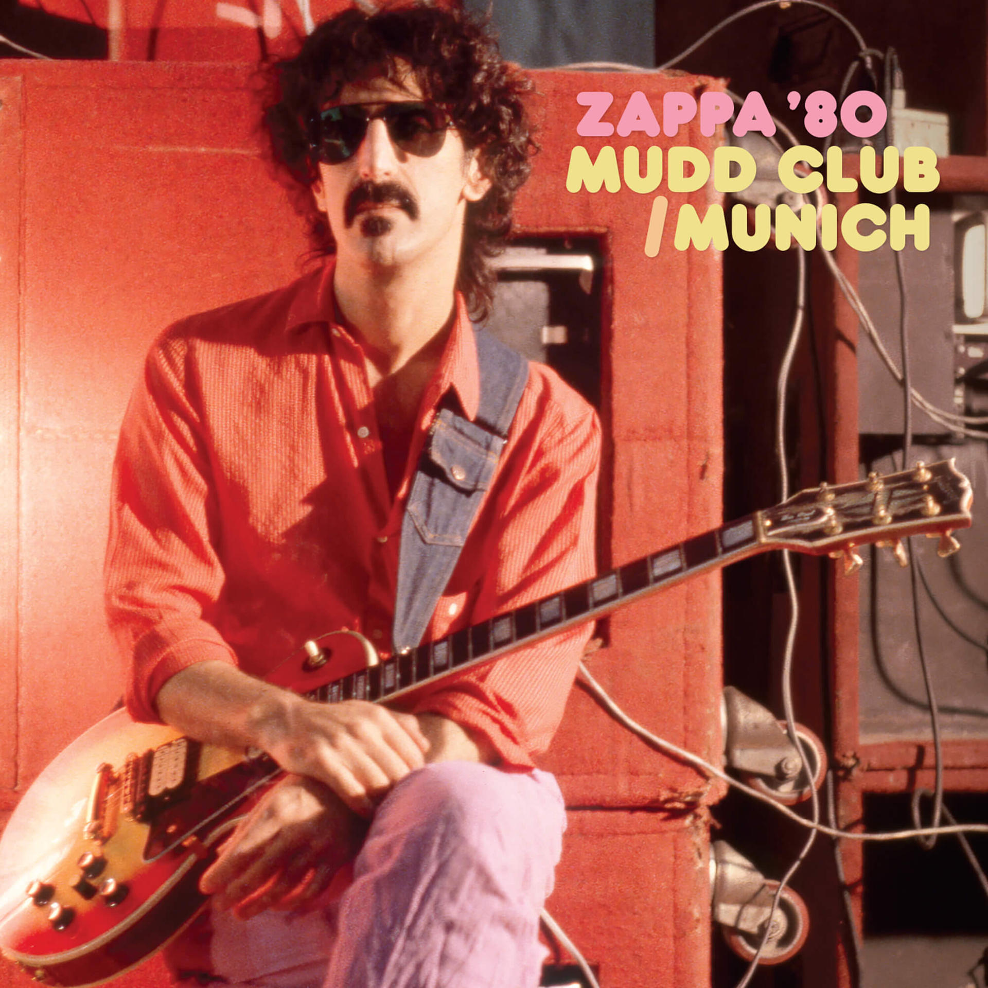 Frank Zappa avec des lunettes de soleil et une moustache, tenant une guitare. Il est devant un amplificateur rouge.