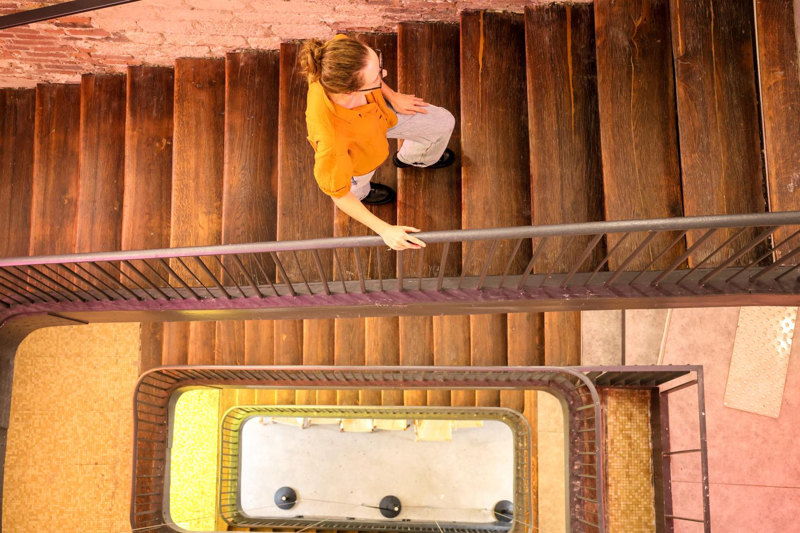 Eine Frau geht Holztreppen hinunter. Die Perspektive ist von oben. Ein Metallgeländer ist sichtbar.
