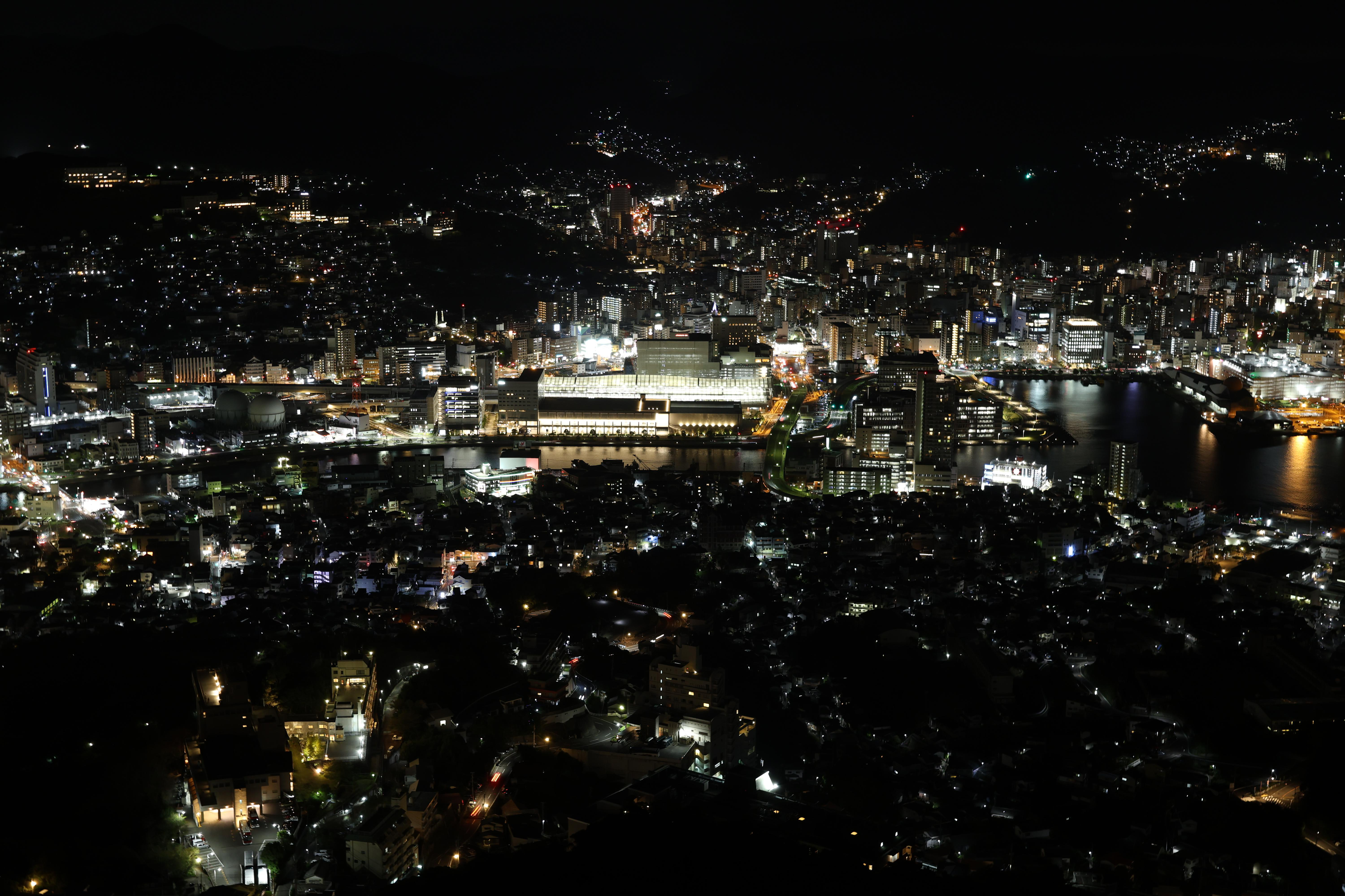 Nachtaufnahme einer Stadt, Lichter beleuchten Gebäude, Straßen und Wasser vor dunklem Himmel.