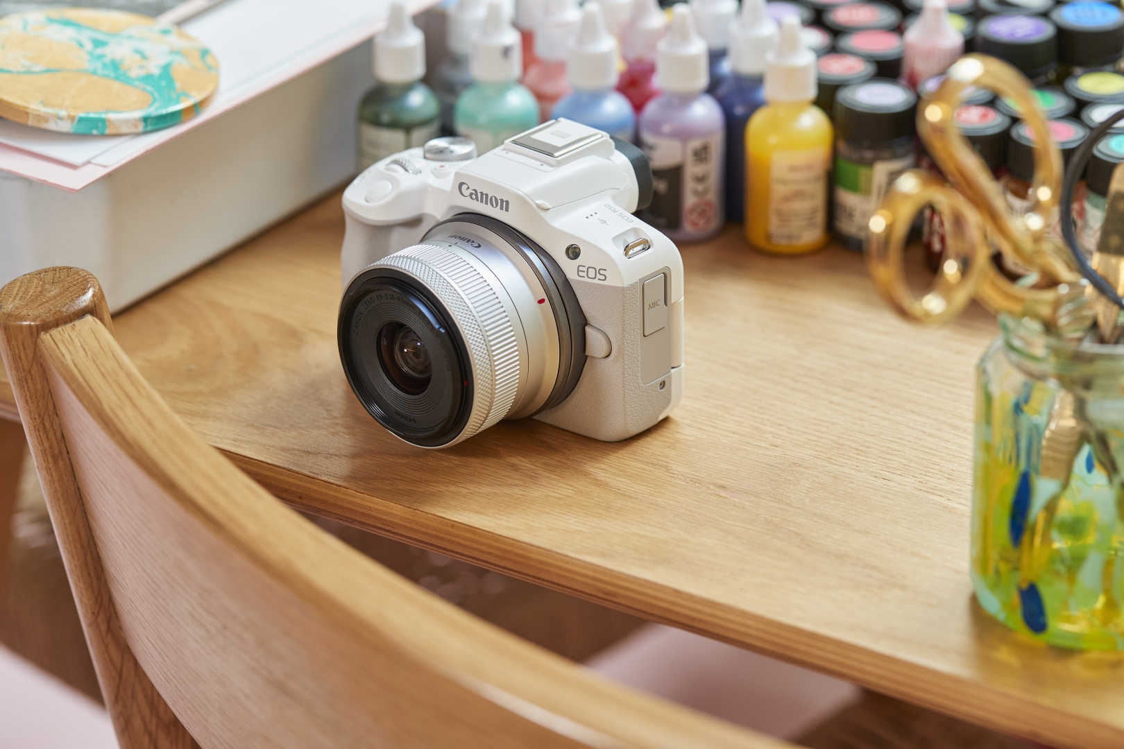 Weiße Canon EOS Kamera auf einem Holztisch, umgeben von Kunstzubehör.