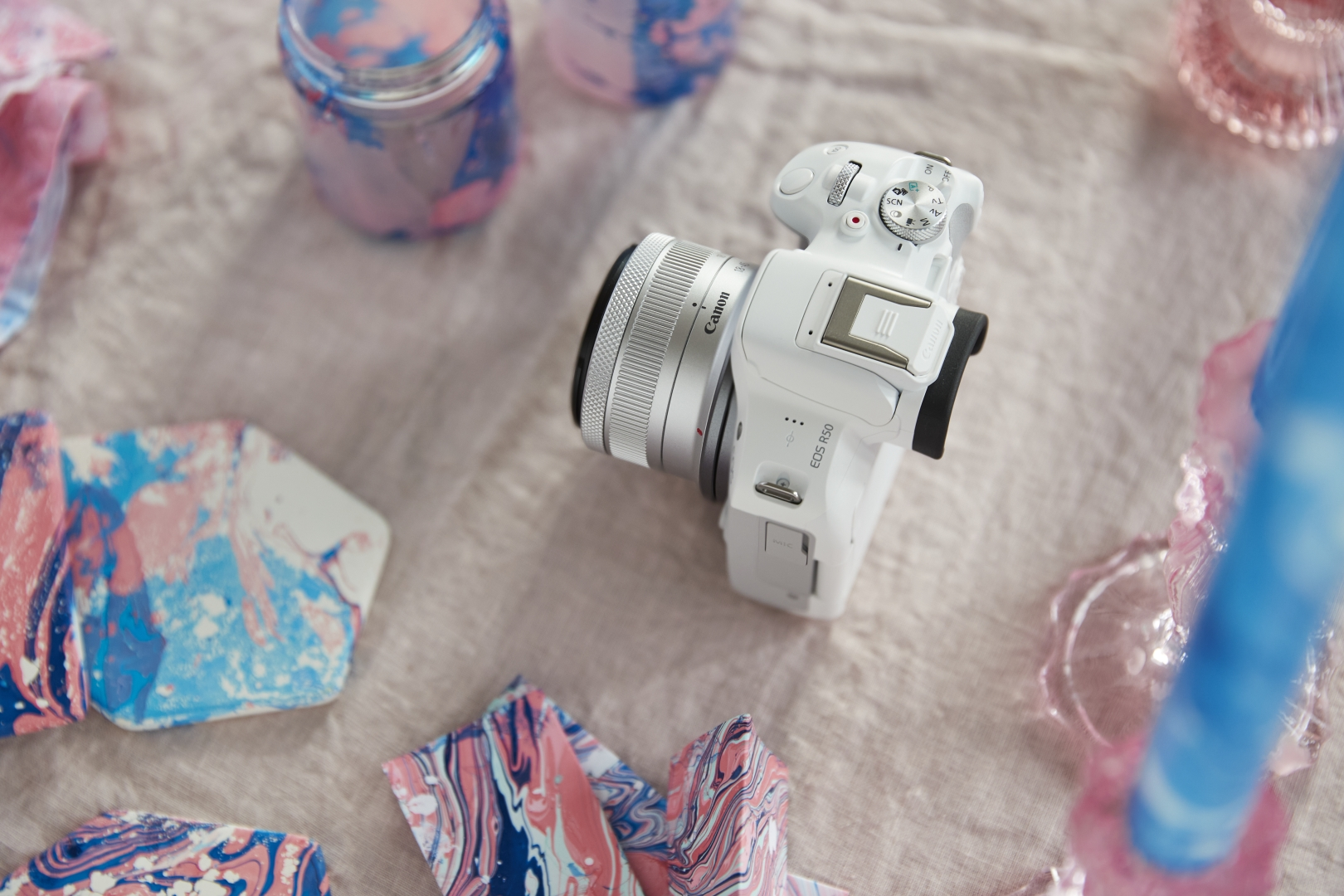 Weiße Canon Kamera mit Objektiv auf einem rosa Tisch, umgeben von Kunstbedarf.