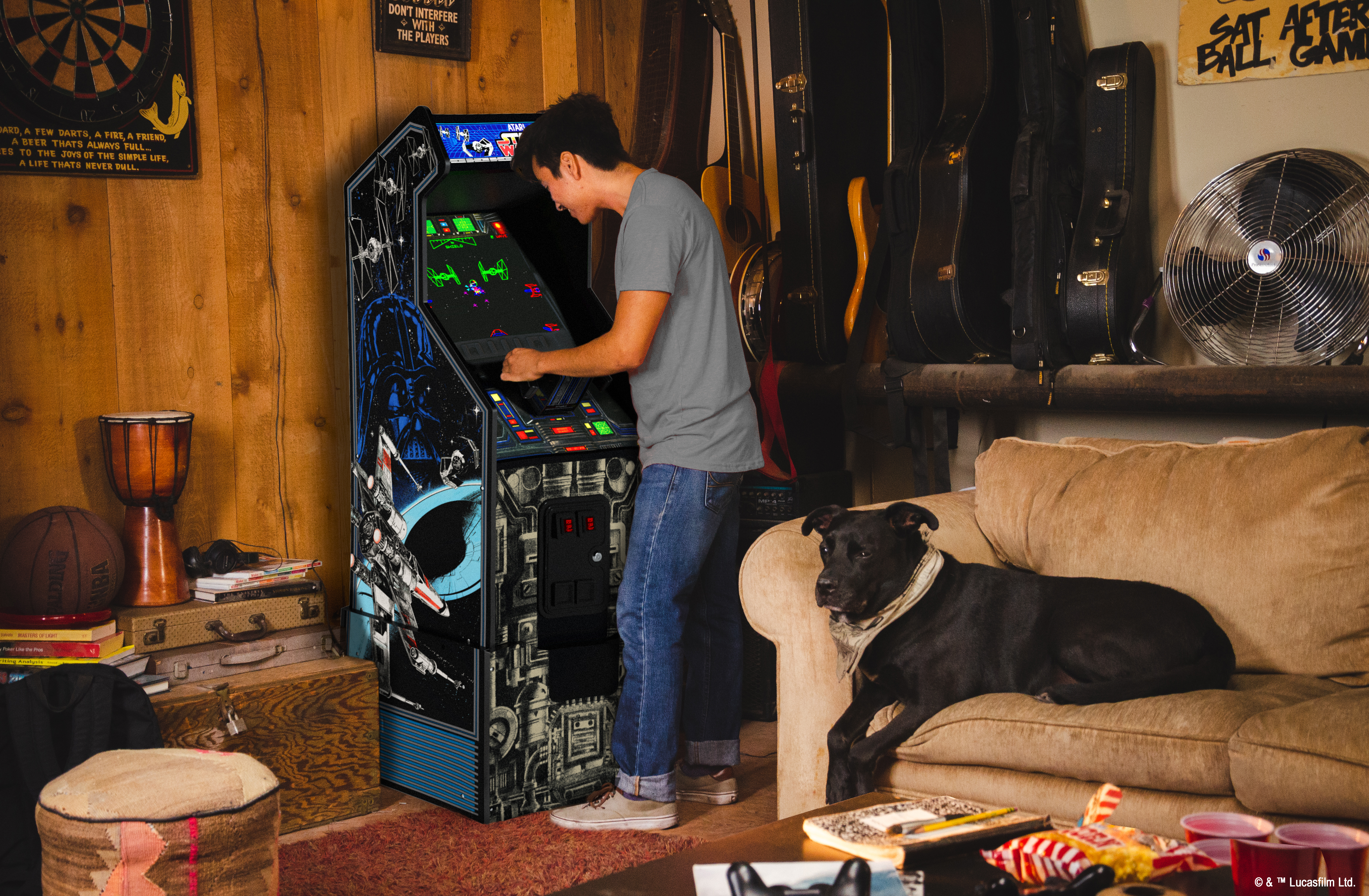 Mann spielt Star Wars Arcade-Spiel, Hund auf Couch, Gitarren an der Wand.